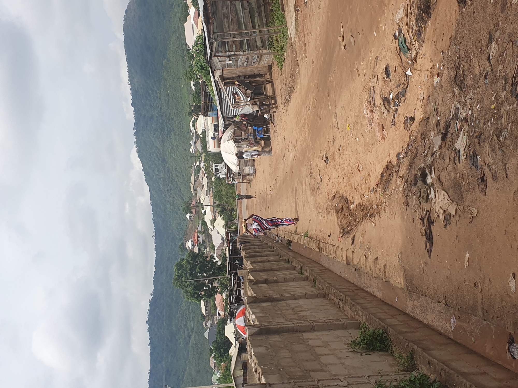 The image depicts a street scene in Lokoja, Nigeria. The unpaved street is lined with buildings, some of which appear to be small shops or stalls. Debris and litter are scattered along the roadside. In the background, a hill or mountain is visible under a cloudy sky. A person wearing a patterned outfit is walking on the stairs running alongside the road. A stone barrier separates the steps and the unpaved road. A second person is walking further up the road, between the stone barrier and the buildings. The image suggests a typical day in a somewhat impoverished area of the city.