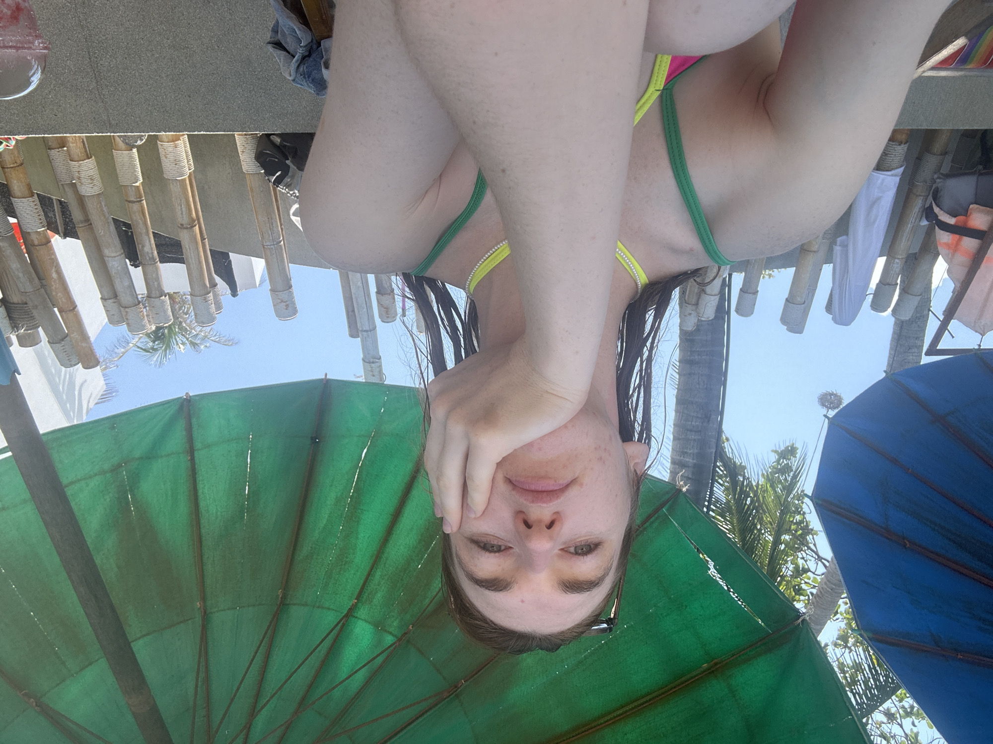 A person, viewed from an upside-down perspective, is situated beneath a green beach umbrella. The person is wearing a bikini top and has wet hair. Their hand is resting on their face. In the background, bamboo structures are visible, along with a clear blue sky and palm trees. To the right, a blue umbrella is present. Based on the location, this is likely the city of Baan Chaweng, Thailand.