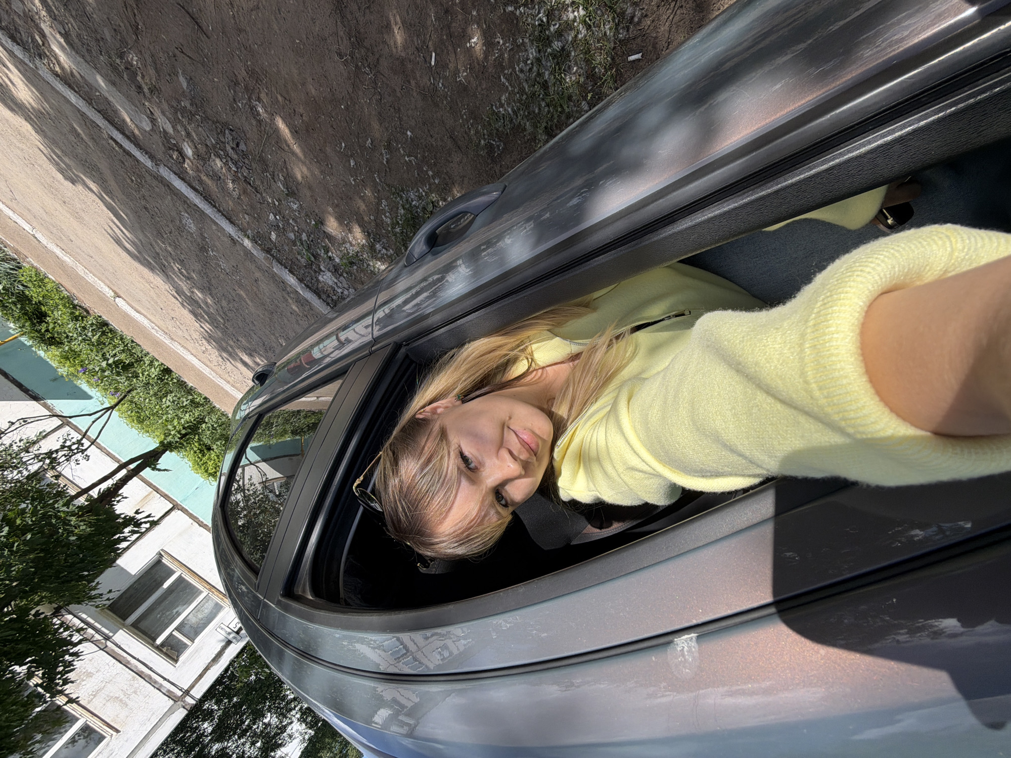 A young woman is taking a selfie from inside a car, leaning out of the open driver's side window. She has blonde hair with bangs, fair skin, and is wearing a light yellow sweater. The car is a silver or gray sedan. Outside the car, there is a building with white walls and several windows. Green trees and bushes are visible near the building. Across the street, there is a path or sidewalk with dirt and some vegetation. Based on the style of the building and environment, the location is likely Chaikovsky, Russia.