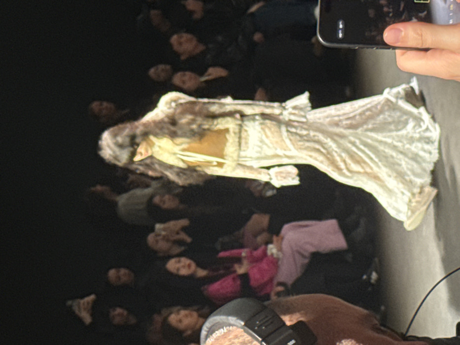 This image captures an indoor fashion show on a dark runway, likely within the city of Paris, France. A female model is prominently featured in the center, walking from left to right. She wears a long, light-colored, semi-sheer lace dress with ruffled cuffs and a ruffled hem, paired with a cropped, tan-brown vest or jacket featuring a light-colored, possibly faux fur, collar. Her long, dark hair obscures her face.
In the blurred background, multiple rows of spectators are seated, observing the event. Several audience members are seen holding up smartphones, actively recording or photographing the runway. One spectator in the mid-ground left is visible wearing a bright pink top and light purple pants.
In the extreme left foreground, the shoulder and part of the head of an individual are visible, wearing black over-ear headphones. A camera flash unit is partially discernible near this individual. In the extreme right foreground, a hand holds a dark-colored smartphone, oriented to capture the model on the runway. The environment is dimly lit, with spotlights illuminating the runway and the model.