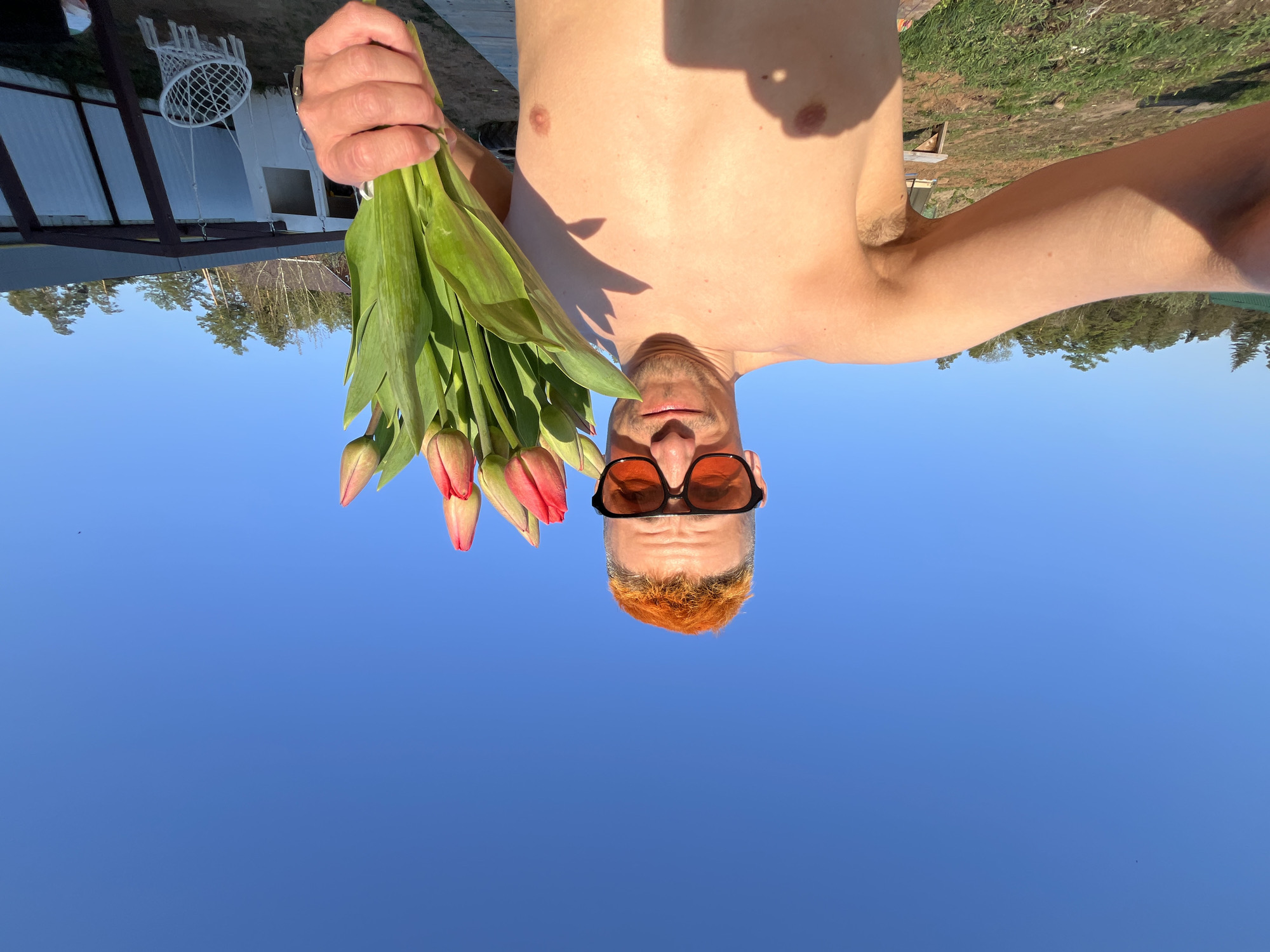 A shirtless man with orange hair, sunglasses, and a fair complexion is holding a bouquet of closed tulips in his right hand. The background is a clear blue sky. Above him, the upside-down view includes trees, part of a white building, and a basketball hoop. The location is Chaikovsky, Russia.