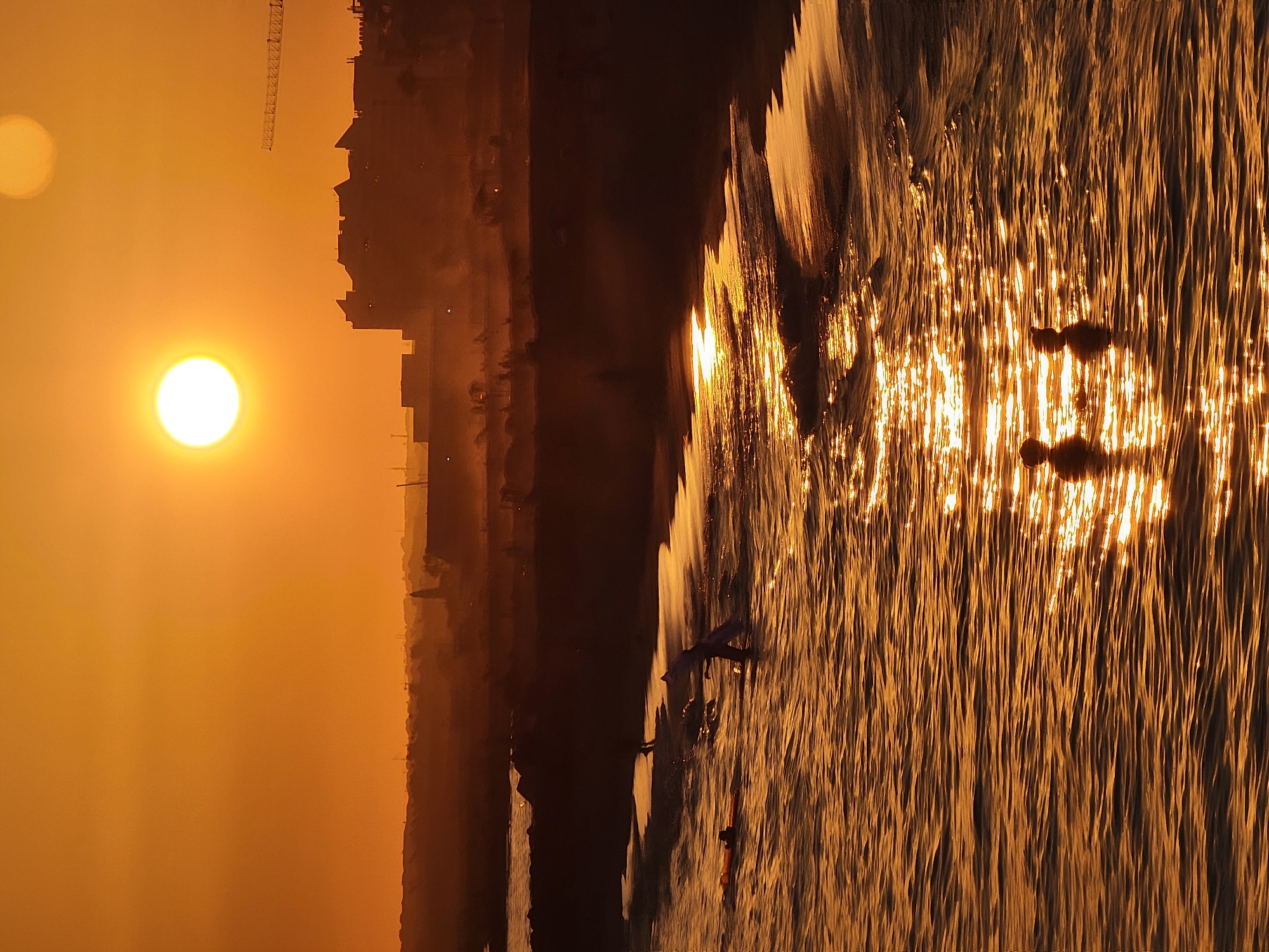 The image captures a sunset over Quarteira, Portugal. The sun is a bright, orb-like presence in the upper portion of the sky, casting a warm, golden hue across the entire scene. In the foreground, the ocean is a shimmering expanse of golden light, with gentle waves lapping at the shore. Two silhouetted figures are visible in the water, their heads and shoulders above the surface. Further out, several other individuals are in the water, some appearing to be swimming or surfing, with a person on a surfboard clearly visible. The shoreline is a dark, silhouetted band, with the faint outlines of buildings and structures visible in the hazy distance. A construction crane is also visible against the sunset. The overall atmosphere is serene and peaceful, with the natural beauty of the sunset amplified by the golden light reflecting off the water.