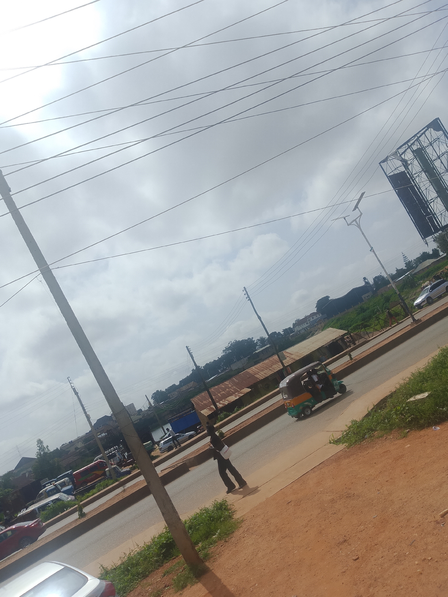 The image depicts a street scene in Bukuru, Nigeria, under an overcast sky. The foreground is dominated by a dirt path and a concrete sidewalk, with lush green grass growing along the edge of the road. A large concrete utility pole, likely for power lines, stands prominently in the middle left of the frame, with numerous wires crisscrossing the sky.
The road is paved and shows signs of traffic. A colorful auto-rickshaw, a common mode of transport in Nigeria, is seen driving away from the viewer, carrying passengers. Further down the road, a silver car is parked or driving. On the right side of the road, a person in dark clothing is walking, carrying a white bag.
In the background, there are various buildings, some with corrugated iron roofs, suggesting a mix of commercial and residential structures. Dense green vegetation covers the rolling hills behind the buildings. To the far right, a large billboard structure is visible against the sky. The overall impression is of a busy, working street in a tropical or sub-tropical urban environment. The time of day appears to be daytime, judging by the natural light. The weather is cloudy, contributing to a soft, diffused light. No discernible text is visible in the image, apart from what might be on the auto-rickshaw, which is not clearly legible.