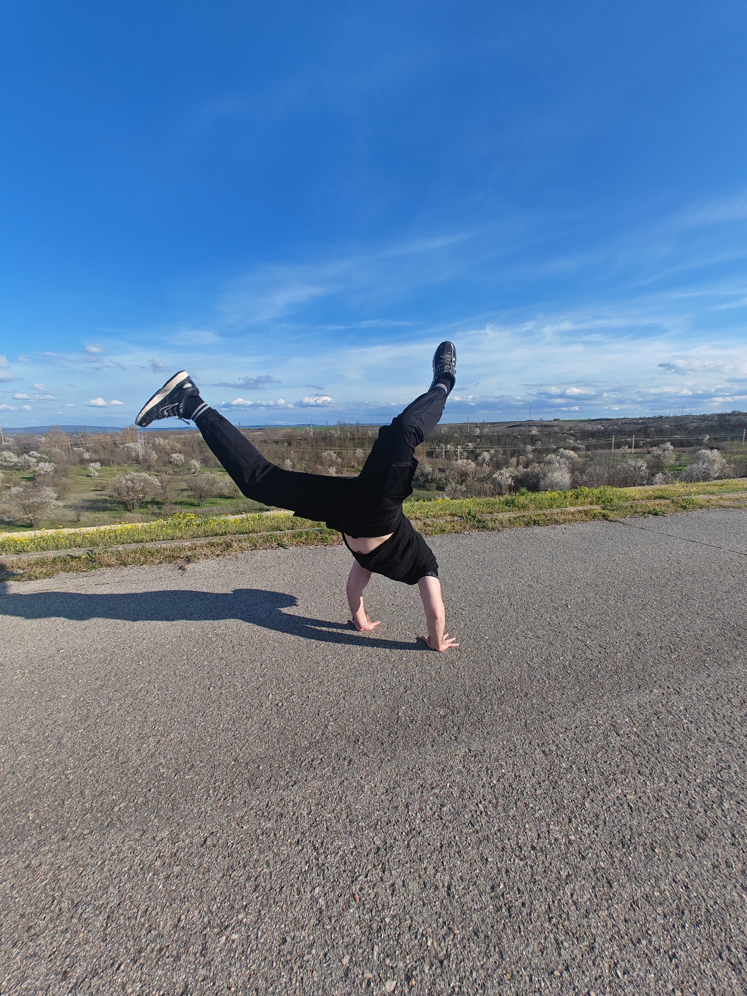A person is performing a handstand on an asphalt surface under a blue sky with scattered clouds. The individual is wearing black pants, a black shirt, and sneakers on one foot. The other foot is bare. The background reveals a landscape with trees and shrubs, suggesting an outdoor location near the city of Shumen, Bulgaria, as inferred from the surrounding environment. The scene appears to be a daytime shot.