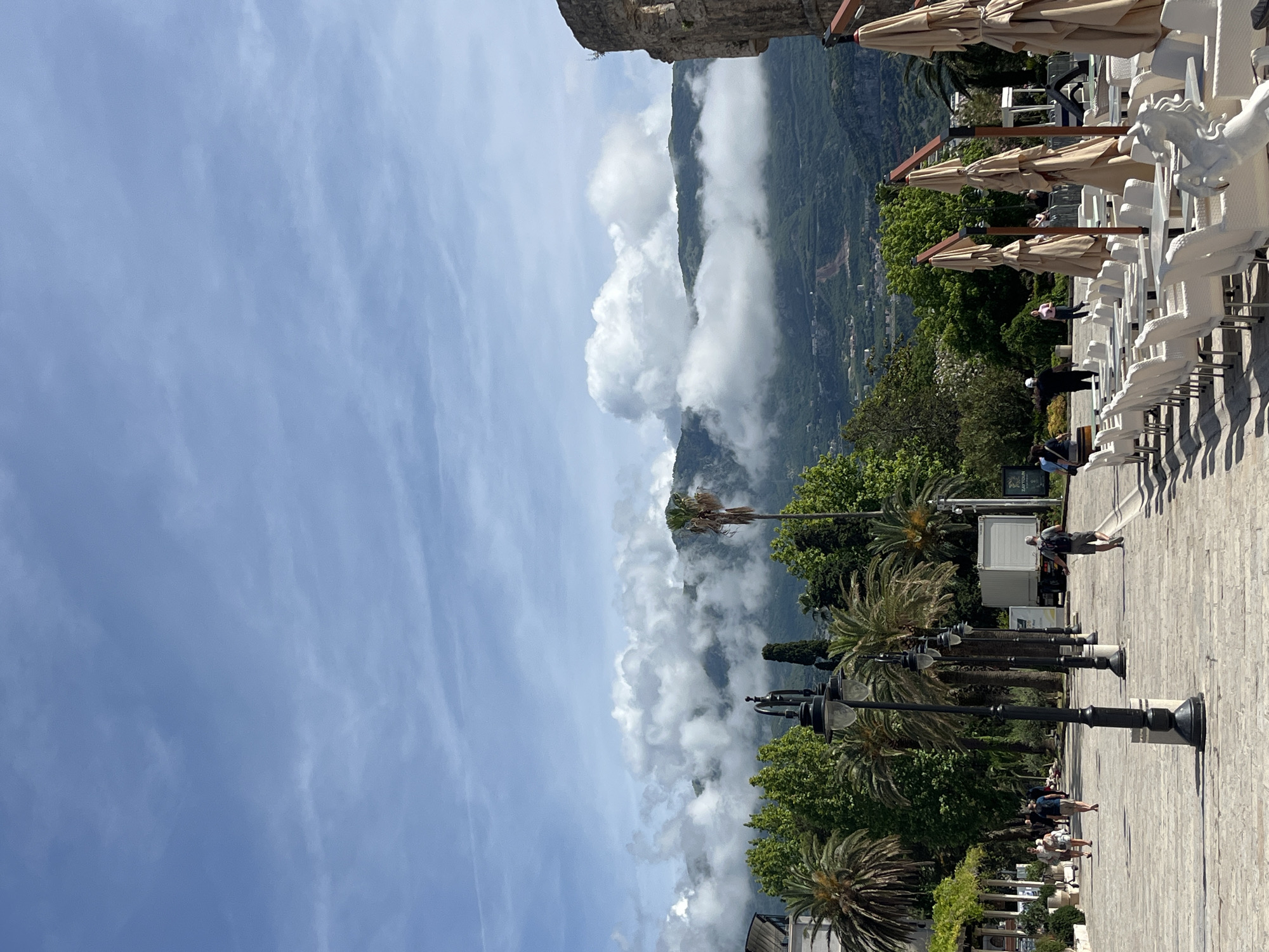 The image captures a panoramic view of a scenic coastal town or area, likely in Budva, Montenegro, overlooking a dramatic mountainous landscape under a bright, partly cloudy sky. The perspective appears to be from a slightly elevated position.
In the background and mid-ground, majestic green mountains are partially shrouded in thick, white, low-lying clouds, creating a misty and atmospheric effect. The mountains are densely forested, with glimpses of a town or settlement nestled in the valleys or on the slopes.
The foreground and mid-ground on the right side of the image feature a wide, paved promenade or plaza made of light-colored stone. Along this promenade, numerous black, ornate streetlights with multiple lanterns are spaced out. To the left of the promenade, there is an outdoor cafe or restaurant area. It is set with many rows of white, wicker-style chairs and beige, folded parasols. Some of the chairs feature distinctive white horse-shaped decorative elements on their backs.
Several people are visible across the scene. Some individuals are walking along the promenade, while others are standing or sitting in the vicinity of the outdoor cafe. Dense green foliage, including tall palm trees and other leafy trees, lines the promenade and adds to the Mediterranean feel of the location.
The sky above is a clear, bright blue, with wispy cirrus clouds stretching across it, complementing the more substantial cumulus clouds clinging to the mountains. The lighting suggests a sunny day, likely midday or early afternoon, as evidenced by the distinct shadows cast by the chairs, parasols, and streetlights. A fragment of an old stone wall or building is visible in the upper left corner. Faint text, possibly "TOURLIN," is visible on a dark sign on the lower right side of the image.