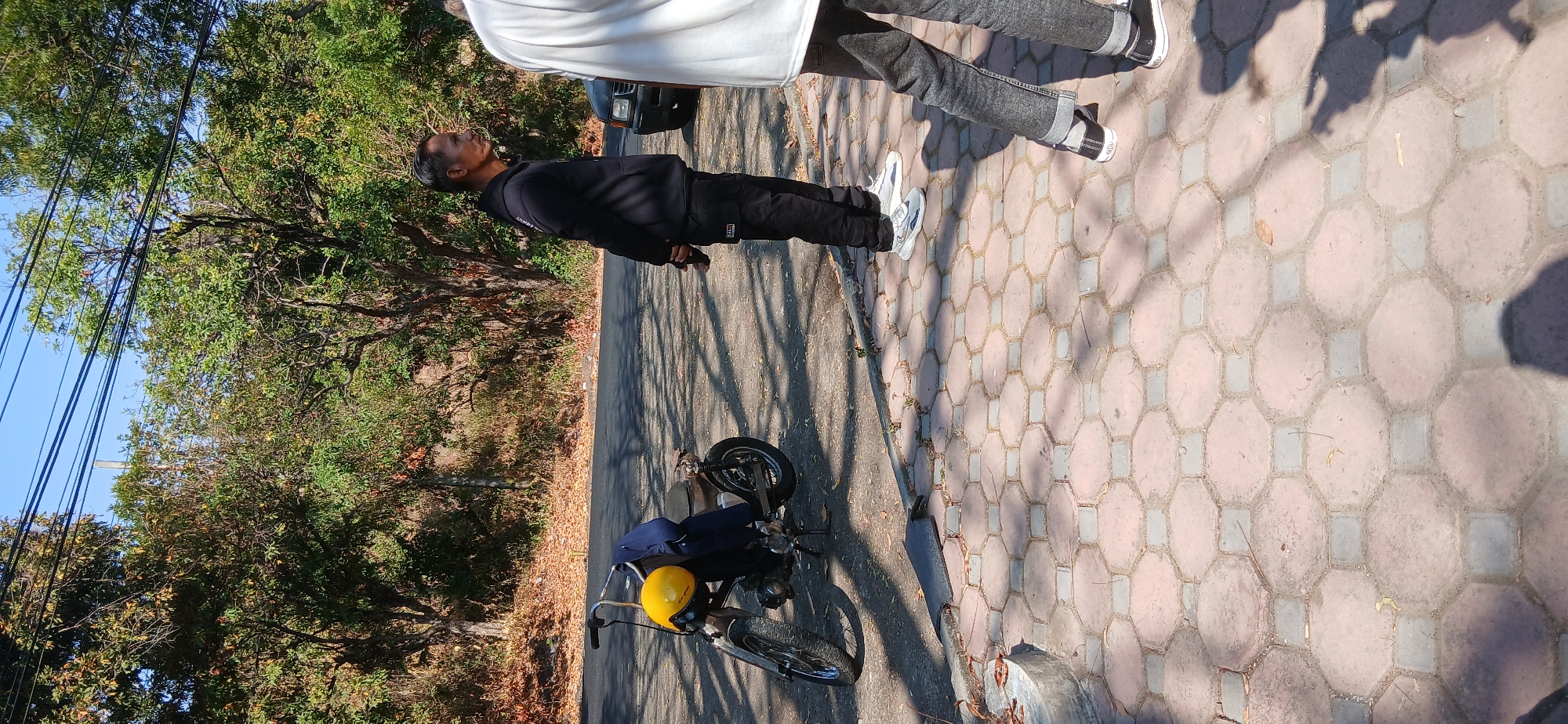 In Banyuglugur, Indonesia, two men are standing near a parked motorcycle on a paved area beside a road. The motorcycle, which appears to be older and has a yellow helmet and a blue garment on its seat, is positioned on the left side of the frame. The man on the right is wearing a black hoodie and black pants, with his hands clasped behind his back. Another person, partially visible from the back, stands to the far right, wearing a white shirt and grey pants. The environment suggests a sunny day, with trees and foliage lining the background and shadows cast on the ground.