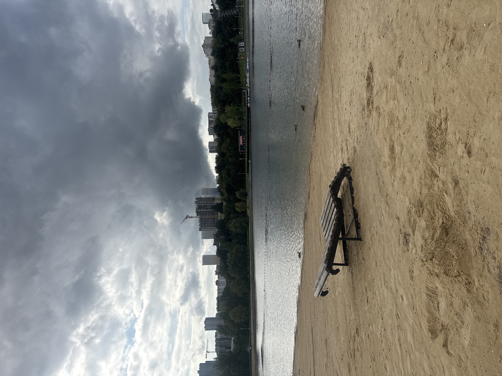 A cloudy day is captured at the Ufa lake, with a sandy beach in the foreground. On the beach, a wooden lounge chair sits empty. The lake stretches out behind the beach, with ducks swimming on its surface. In the background, a line of trees separates the lake from the city skyline, which is dominated by modern buildings and construction cranes. The sky is filled with dark, heavy clouds, suggesting an impending storm.