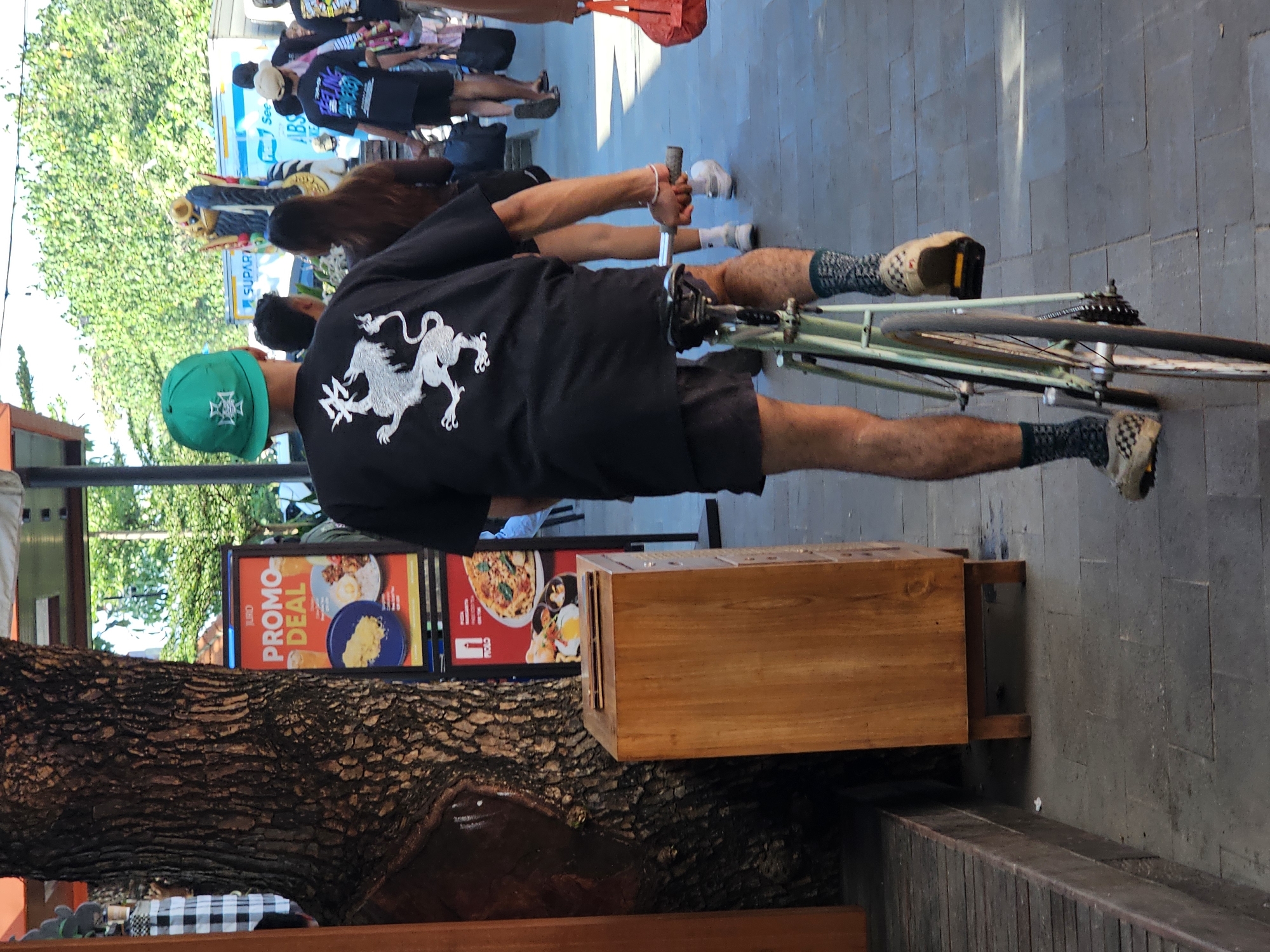 A person is riding a light green bicycle down a paved sidewalk in Denpasar, Indonesia. The rider, viewed from behind, wears a black t-shirt with a white graphic of a lion-like creature, black shorts, patterned socks, and tan checkered slip-on shoes. They also wear a green baseball cap. The bicycle has a silver handlebar and a dark-colored seat. To the left of the rider is a large, rough-barked tree and a tall, wooden trash receptacle. A red and yellow promotional sign with an image of food is visible in the background, along with other pedestrians and buildings. The scene appears to be outdoors during the daytime, with bright sunlight casting shadows.