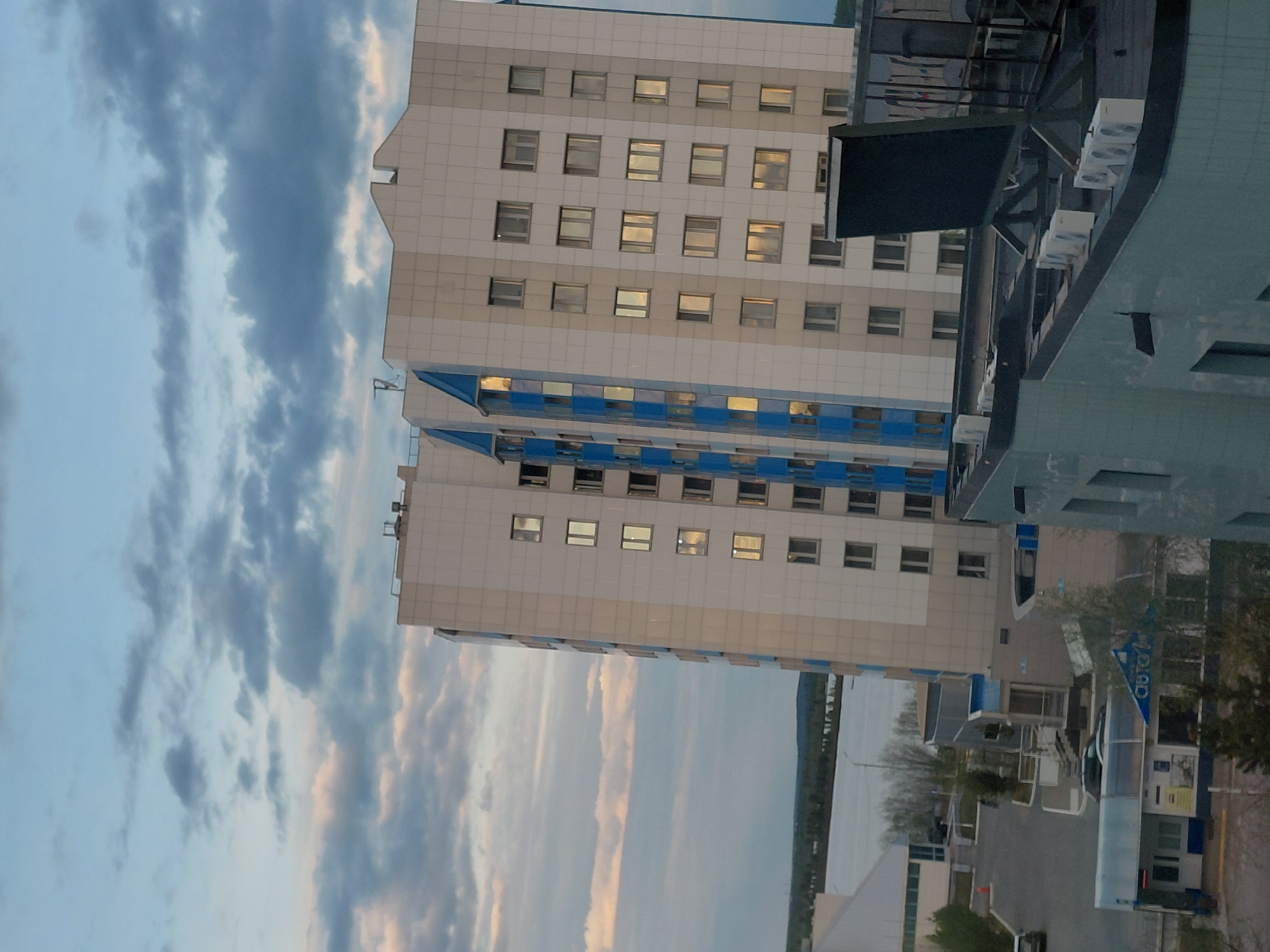 The media file presents an outdoor urban landscape, likely captured from an elevated position, overlooking buildings, a body of water, and the sky. The image appears to be taken during late afternoon or early evening, characterized by soft light and a cloudy sky.
The central subject is a multi-story building with a light-colored (beige or off-white) facade and numerous rectangular windows. The lower sections of this building feature distinctive horizontal blue bands. Many windows on the upper floors show reflections of warm light, possibly from interior illumination or the setting sun. To the right of this building is another, darker structure, possibly made of dark panels or glass, which has a more angular and modern design. This darker building features several white air conditioning units mounted on its exterior, and a large, dark rectangular structure (possibly a screen or sign) is affixed to it.
The sky occupies a significant portion of the scene, filled with a mix of light blue and grey, streaky clouds, suggesting a dynamic atmosphere but not necessarily stormy weather. In the mid-ground, a wide body of water, likely a river or estuary, stretches towards the horizon, with a low landmass visible on the distant shore.
At the ground level, particularly towards the bottom right of the upright image, elements of an urban environment are visible, including roads, some scattered trees, and a commercial-looking structure. On a blue sign associated with this ground-level structure, the Russian text "AВТО" (AVTO), meaning "AUTO," is discernible. No explicit human activity is clearly visible, making the scene appear calm and still. The overall impression is a modern cityscape view at twilight.