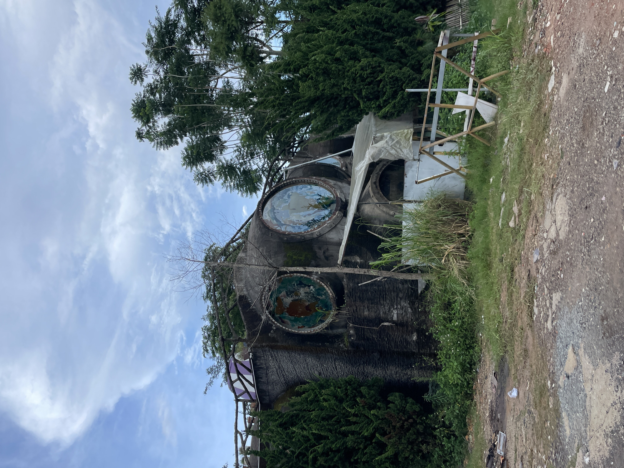 The image depicts an abandoned, somewhat surreal-looking concrete structure with two large, circular openings on its facade, resembling eyes. These openings contain weathered, circular paintings, one with abstract green and brown splotches and the other with a faded depiction of underwater flora and fauna. The structure's exterior is dark and textured, with signs of age and neglect, covered in moss and vines in some areas.
A temporary, makeshift shelter made of translucent plastic sheeting and scaffolding is attached to the front of the building, suggesting some form of ongoing, albeit rudimentary, activity. In front of this shelter, a wooden A-frame structure, possibly a display or support, stands with a white panel propped within it.
The building is surrounded by dense vegetation, including tall cypress-like trees on the right and various grasses and plants at its base. The ground in the foreground is a mix of rough dirt, gravel, and patches of worn concrete, littered with debris, including plastic bottles and discarded materials, indicating a disused or construction site. The sky above is partly cloudy with scattered white clouds against a pale blue background.
This structure is located in Da Lat, Vietnam, a city known for its unique architecture, often featuring French colonial influences and avant-garde designs. While the specific name or purpose of this particular building isn't evident from the image alone, its eccentric design and isolated, somewhat overgrown setting are characteristic of some of the less-maintained or abandoned artistic installations and unconventional structures found in and around Da Lat. It evokes a sense of mystery and forgotten artistic endeavor.