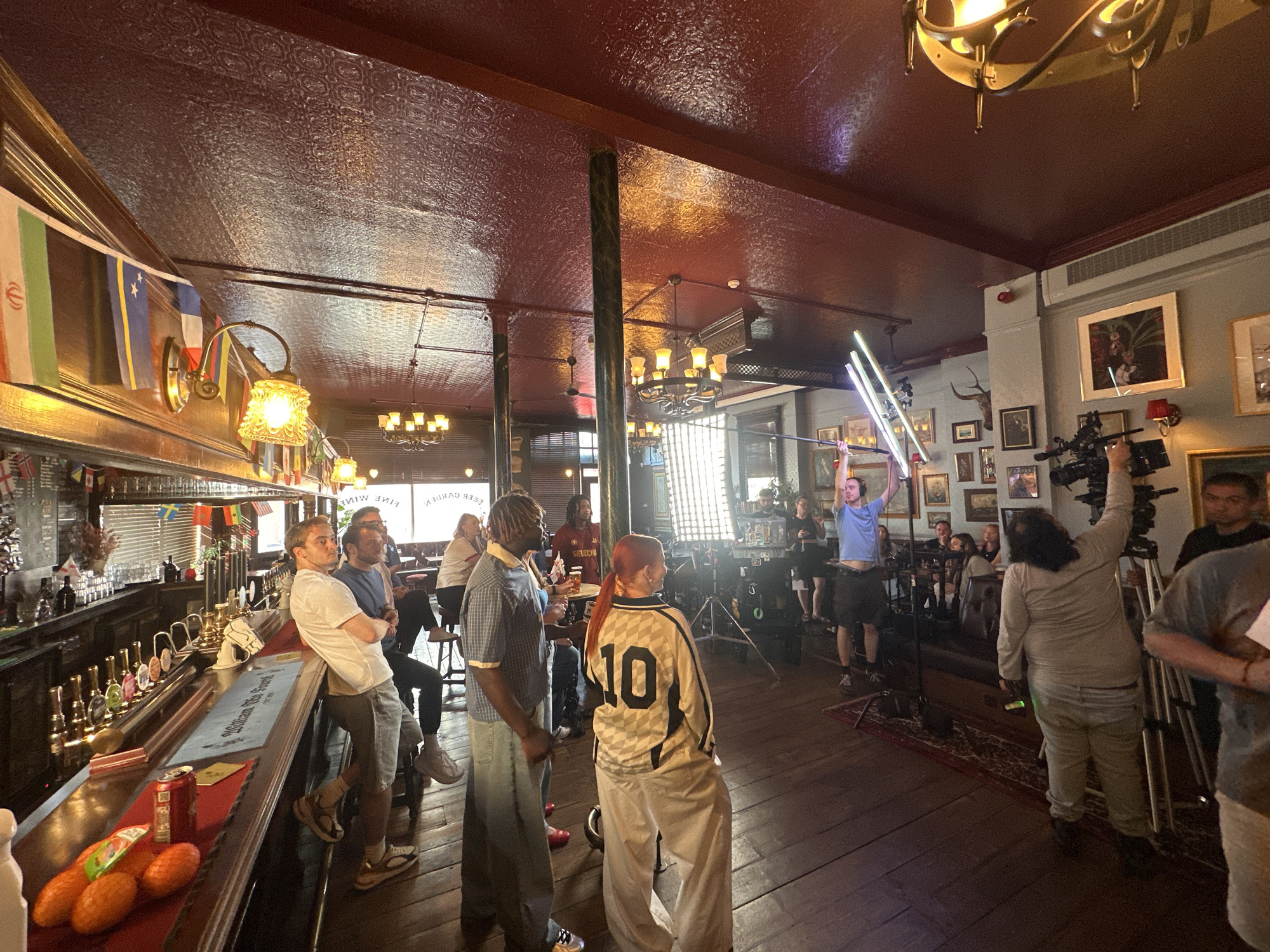 The image captures a scene inside a traditional British pub, possibly located in London, judging by the architectural style and atmosphere. The room is crowded with people, suggesting it could be a busy afternoon or evening.
On the left, several individuals are standing or leaning against a bar, which is adorned with a variety of drink taps, bottles, and what appear to be decorative fruits. A can is placed on the bar. Flags of various nations hang from the ceiling above the bar.
The center of the image is occupied by people engaged in what seems to be a film or video shoot. There is camera equipment present, including a camera and a lighting setup held by a person in the background. Other people are standing around, potentially part of the crew or cast. One person, wearing a shirt with the number "10" on it, is in the foreground.
The environment is warm and dimly lit, with dark wood flooring and a decorative patterned ceiling. The walls are covered with various framed pictures and mounted animal heads, adding to the traditional pub decor. Chandeliers hang from the ceiling, contributing to the old-world charm of the location. The floor is made of dark planks.