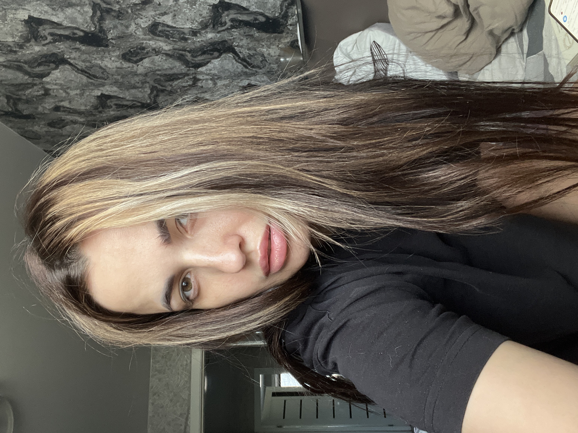 A young woman with long, highlighted brown and blonde hair is centered in the frame, looking directly at the camera. She is wearing a black, long-sleeved shirt. The background includes a marble-patterned wall behind her and a bed with beige and white linens in the upper right. A white radiator can be seen in the lower center of the frame. Location is not specified.