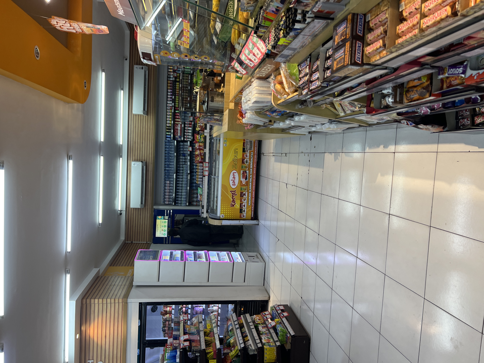 The image captures the interior of a convenience store, possibly a mini-mart or a minimarket, located in Denpasar, Indonesia. The store is well-lit with fluorescent ceiling lights, and the floor is covered with white tiles. The perspective is from the entrance area, looking deeper into the store.
On the left side of the frame, a tall refrigerator unit filled with various products is visible. Next to it stands a white display unit with multiple shelves illuminated by pink neon lights, likely for showcasing specific items or promotions. Further down this aisle, shelves are stocked with packaged goods, including snacks and beverages.
In the center of the image, a refrigerated display case for food products, possibly deli items or pre-packaged meals, is prominent. It features yellow signage with text that appears to be in Indonesian, "Roti Salam Zaitun" (Bread Olive Salam) and "MAKANAN OLAHAN HALAL" (Halal Processed Food). Two individuals, dressed in dark clothing, are standing near this display, seemingly customers or staff. One appears to be facing away from the camera.
To the right, a wide variety of products are displayed on shelves, including candies, snacks, and other convenience items. A prominent "BAKERY" sign is visible, indicating a section dedicated to baked goods. The shelves are densely packed with colorful packaging. A glimpse of a checkout counter or service area can be seen in the background, along with more shelving and a large display of bottled drinks. The overall impression is that of a bustling retail environment. The distinct orange ceiling detail further adds to the store's aesthetic.