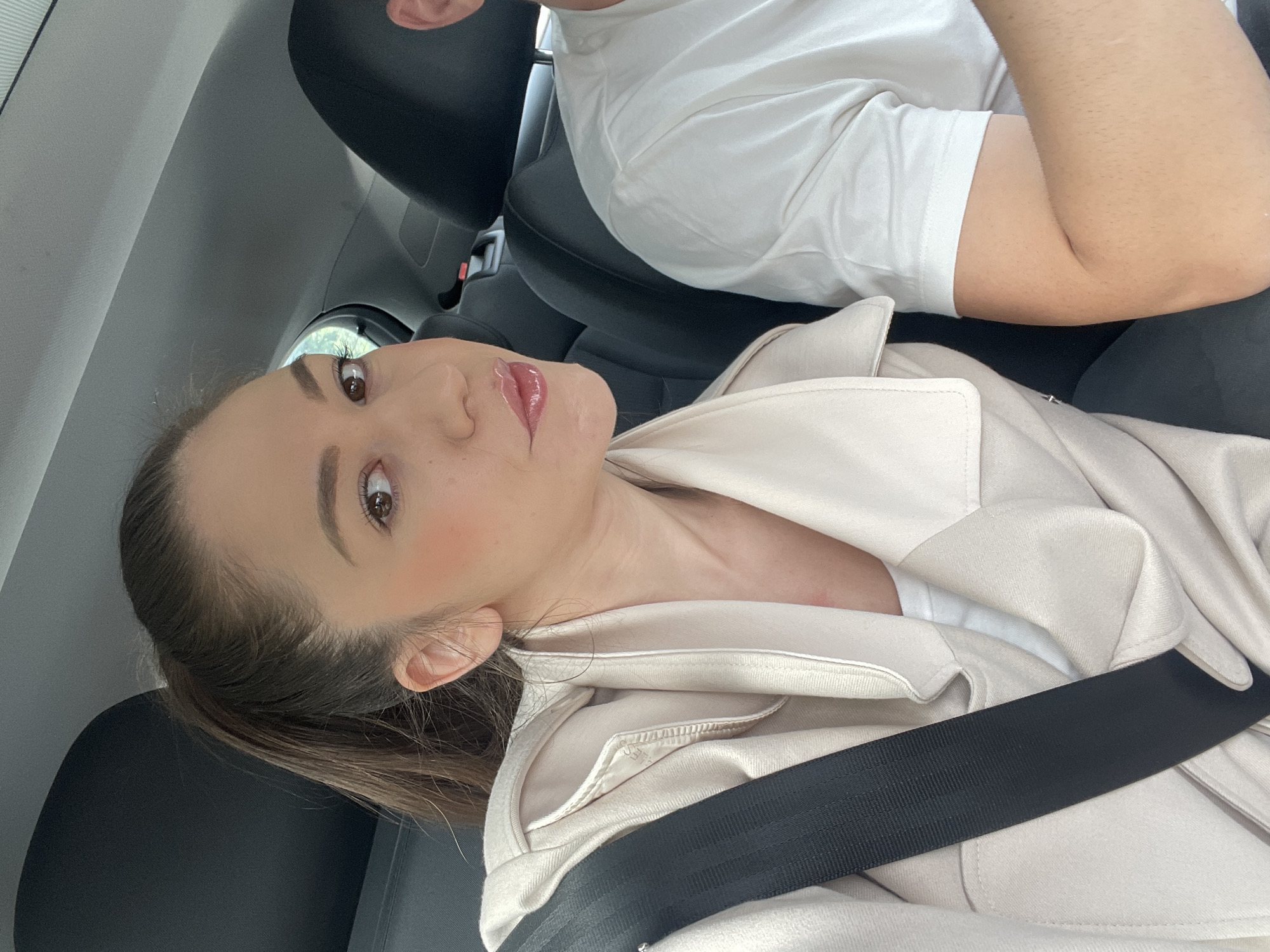 A selfie-style image captures two individuals inside a car during daylight hours. The primary subject, a young woman, is in the foreground, facing the camera with a direct gaze and a slight smirk. She has brown hair tied back and is wearing a light beige or cream-colored jacket over a white top, secured by a black seatbelt across her chest. Her makeup includes defined eyebrows, mascara, and glossy pink lips.
Partially visible behind her is a second person, likely male, wearing a white short-sleeved t-shirt. Only his upper body and arm are seen, suggesting he is seated in the front passenger or driver's seat.
The car's interior features dark seating and light grey ceiling upholstery. The bright lighting indicates it is daytime. No specific landmarks or textual elements are visible to provide a precise location, beyond the general setting of a vehicle. The atmosphere is casual, portraying a moment during travel.