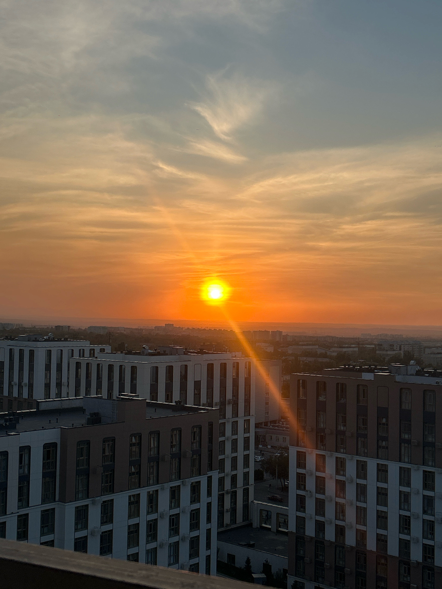 The image shows a panoramic view of a city during sunset. The sky is filled with warm orange and yellow hues, with some wispy clouds. The sun is a bright, intense orb near the horizon, casting a strong ray of light across the scene. In the foreground, there are several modern, multi-story apartment buildings with a consistent architectural style, featuring white and brown facades and numerous windows. These buildings are arranged in rows, suggesting a planned urban development. Beyond the buildings, the cityscape stretches into the distance, with more structures and hints of green trees visible. The overall impression is that of an urban landscape bathed in the soft, warm light of a setting sun. The scene appears to be a residential area with high-density housing. The angle of the shot suggests it was taken from a higher vantage point, possibly another building or a balcony.