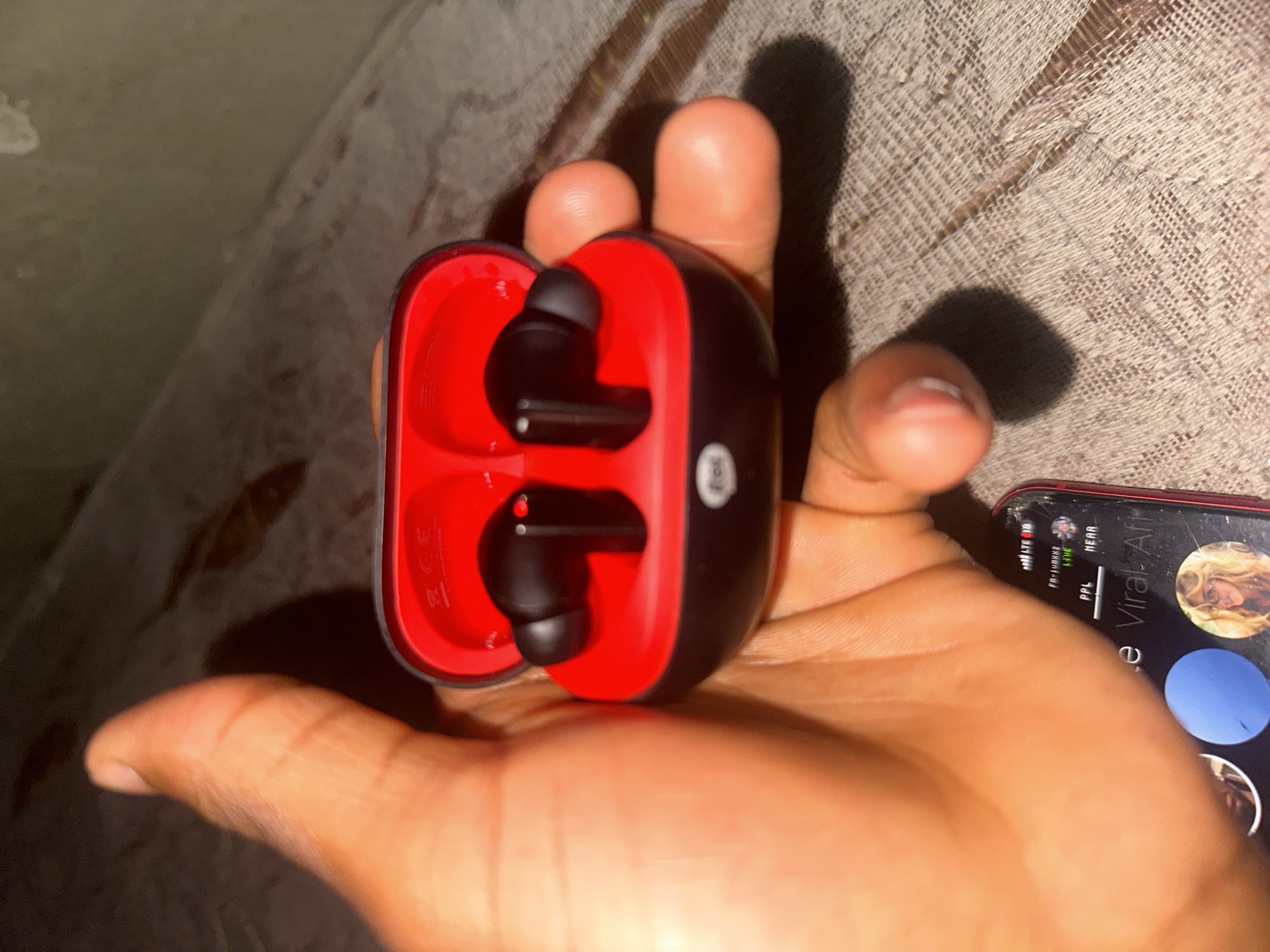 An overhead shot captures a hand holding a black and red wireless earbud charging case open, revealing two black earbuds inside. The case rests on a textured, light brown surface, possibly a blanket or fabric. A smartphone is visible in the lower portion of the image, its screen displaying notifications and profile pictures. The image appears to be taken indoors.
