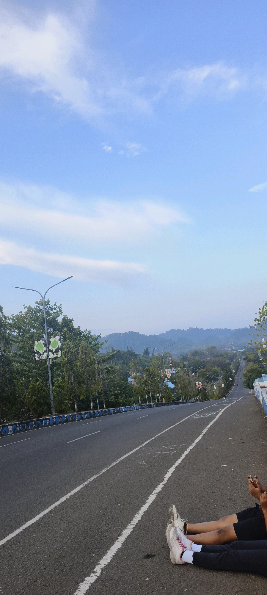The image depicts a road in Waisai, Indonesia, on a clear day with a blue sky and scattered clouds. The road appears to be a well-maintained asphalt surface with clear white lane markings. On the left side of the road, there are trees and streetlights, some of which are decorated with graphics of turtles. A decorative blue and white barrier runs along the side of the road. In the background, lush green hills rise, shrouded in a slight mist.
In the foreground, on the right side of the road, two individuals are seen sitting on the asphalt. Their legs are extended, and they are both wearing athletic shoes. One person is wearing white shoes with pink laces, and the other is wearing white shoes with white laces. Their clothing consists of dark-colored pants and shorts. They appear to be resting or taking a break. The overall impression is a serene outdoor scene, possibly a scenic viewpoint or a road leading into a natural landscape.