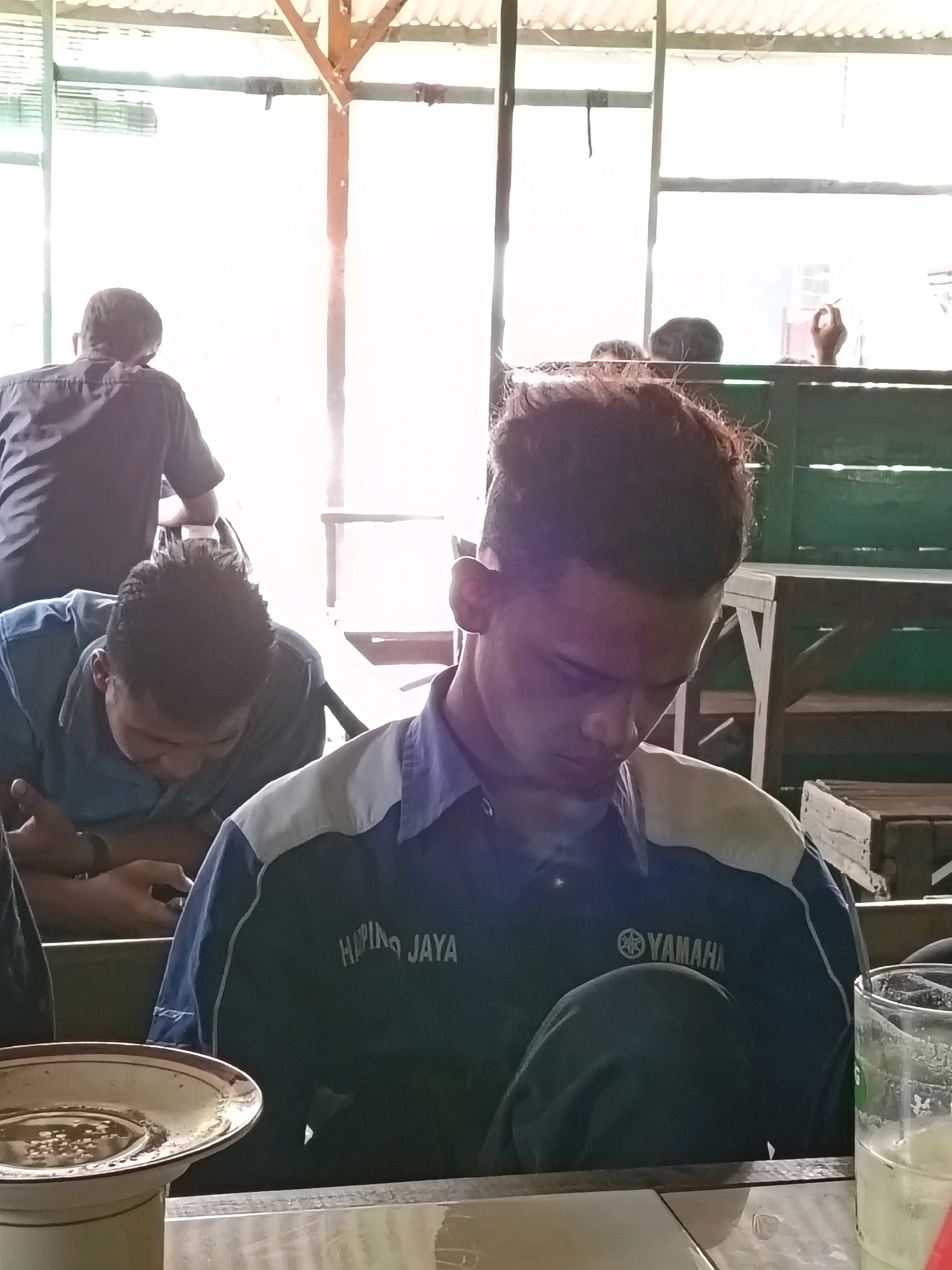 The image depicts a group of men in what appears to be an open-air seating area, possibly a roadside food stall or a casual dining spot, in Blora, Indonesia. In the foreground, a young man with styled hair is seated, looking downwards, wearing a dark blue polo shirt with white and light blue accents on the shoulders and sleeves. The shirt has visible text that reads "HAPING JAYA" and the Yamaha logo. Next to him, on his right, is a partially visible glass with a light green liquid, likely a beverage. To the left of the foreground subject, another man in a blue shirt is also looking down, possibly at a mobile phone. In the background, another man is visible from the back, wearing a dark collared shirt. The environment features wooden structures, possibly supporting a corrugated metal roof, and green wooden benches or tables. The lighting suggests it is daytime, with sunlight filtering through the roof, creating a bright, somewhat washed-out effect. The overall scene conveys a casual, everyday setting in a local Indonesian community.