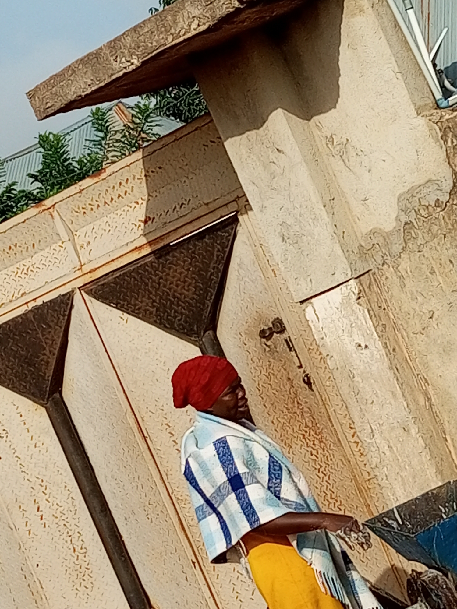 A person wearing a red headscarf and a blue and white checkered shawl stands near a metallic gate in Gombe, Nigeria. The person is also wearing a yellow garment. The background consists of textured walls, possibly part of a building or a boundary, with some vegetation visible above a corrugated metal roof. The overall scene appears to be outdoors, under daylight.