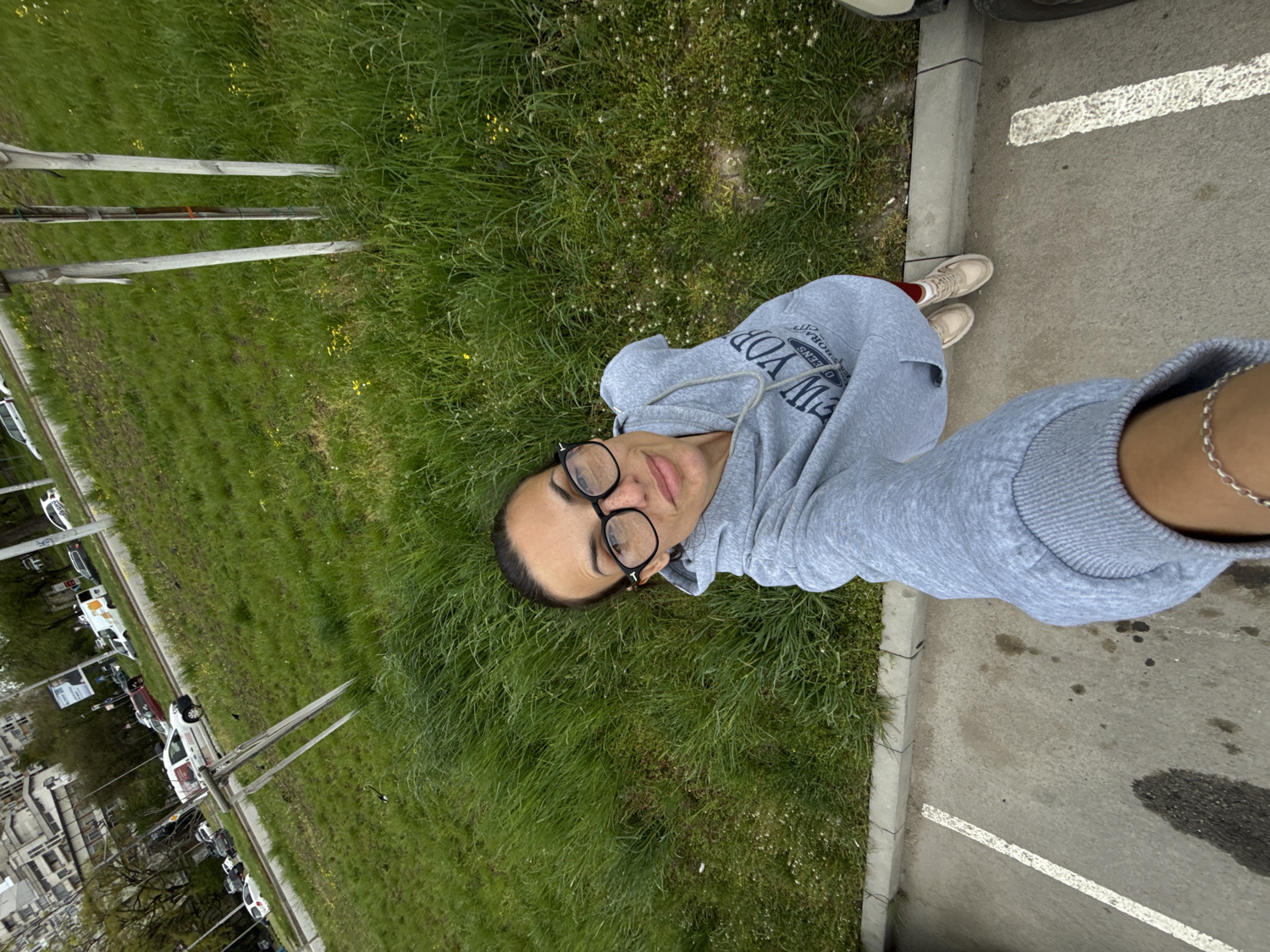The image is a vertical selfie captured outdoors, featuring a woman from the chest up, looking directly at the camera. She wears black-rimmed glasses and has dark hair pulled back. Her expression is neutral, with a slight, subtle smile. She is dressed in a light grey hooded sweatshirt (hoodie) which has white text on the front, partially visible, reading "NEW YORK," "QUEENS," and "ORIGINAL." A silver chain bracelet is visible on her right wrist.
The woman is situated at the edge of a vibrant green grassy area, which occupies the left side of the frame. To her right is a grey paved surface, likely a sidewalk or a parking lot, marked with a white painted line. In the background, beyond the grassy strip and along what appears to be a street, there are several parked cars, utility poles, and parts of buildings. The lighting is soft and even, suggesting it is daytime, possibly under an overcast sky. The overall scene depicts an urban or suburban environment in Pleven, Bulgaria.