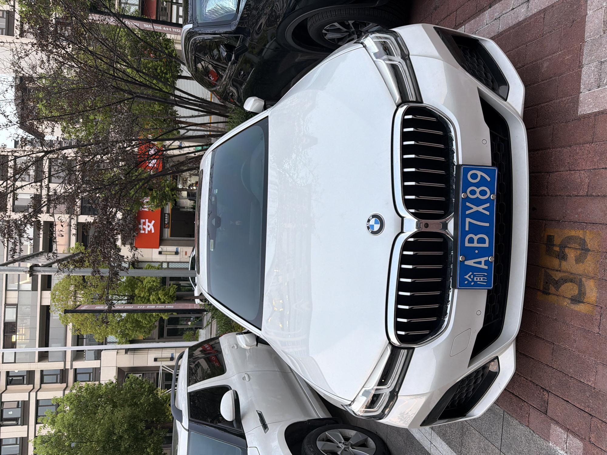 A white BMW sedan is parked on a brick-patterned ground in a city. The car has a prominent grille and the BMW logo on the front. A blue license plate is affixed to the front bumper, displaying the characters "渝A·B7X89". To the left of the BMW, a white SUV is partially visible. To the right, a dark-colored SUV is parked. In the background, a multi-story building with numerous windows is visible, along with trees and an orange sign with Chinese characters. The scene appears to be an urban parking lot.