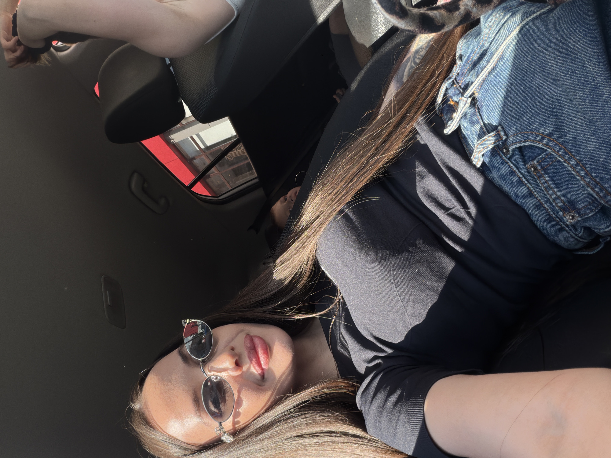 The image, rotated 90 degrees clockwise, is a sunlit selfie taken inside a car, likely in Izhevsk, Russia. The primary subject is a young woman, seen from a low angle, looking upwards towards the camera. She wears a black long-sleeved top and reflective, round sunglasses. Her long, straight brown hair is prominent, illuminated by direct sunlight that casts strong shadows across her face and clothing. She has her tongue playfully extended between her lips.
A blue denim garment, possibly jeans or a jacket, is visible to her right, along with a blurred, patterned object that resembles a cat's tail or an animal print fabric. To her left, the arm and shoulder of another person, clad in light-colored clothing, are visible in the foreground. Further back in the car, a bare foot can be seen on the dark floor, indicating another passenger.
The car's interior features dark seats, a headrest, and a grab handle on the ceiling. Through the car windows and reflected in the sunglasses, exterior structures with distinct red and white elements are visible, suggesting an urban or commercial environment. The bright lighting indicates it is daytime, likely a sunny day. The overall atmosphere is casual and relaxed. No visible text is present in the image.