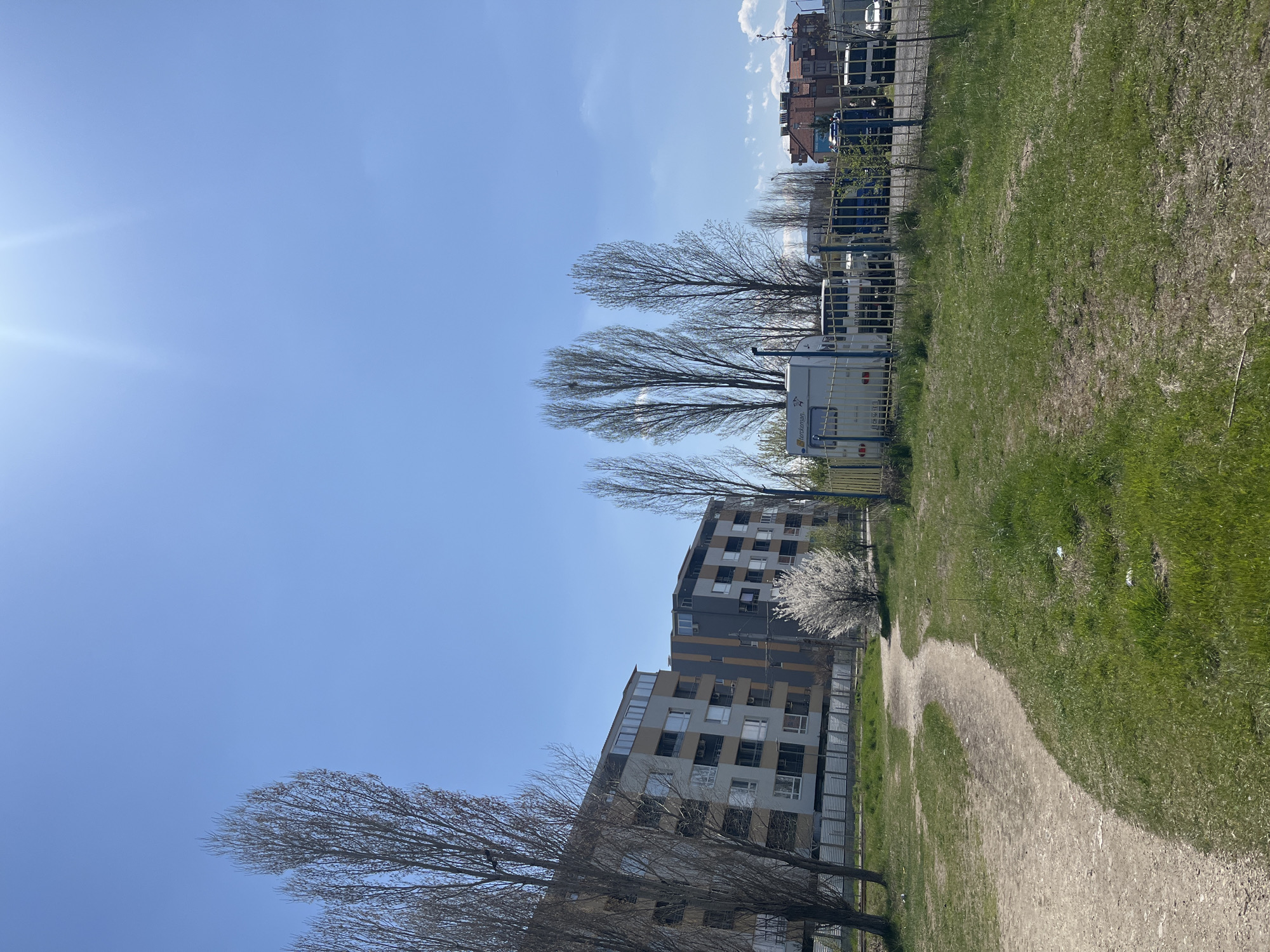 The image depicts an outdoor scene in Sofia, Bulgaria, characterized by a mix of urban and natural elements. In the foreground is a dirt path meandering through a grassy area. To the right of the path is a patch of green grass. Adjacent to the grassy area is a fenced enclosure holding what appear to be recreational vehicles, trailers, and a small utility vehicle. The fence lines the edge of the park. Beyond these, several trees with bare branches are visible, hinting at early spring. Further back, a modern apartment building rises, its façade a mix of gray, tan, and white sections. In the distance, there are more buildings, but they are less distinct. The sky is clear and blue, with the sun shining brightly.