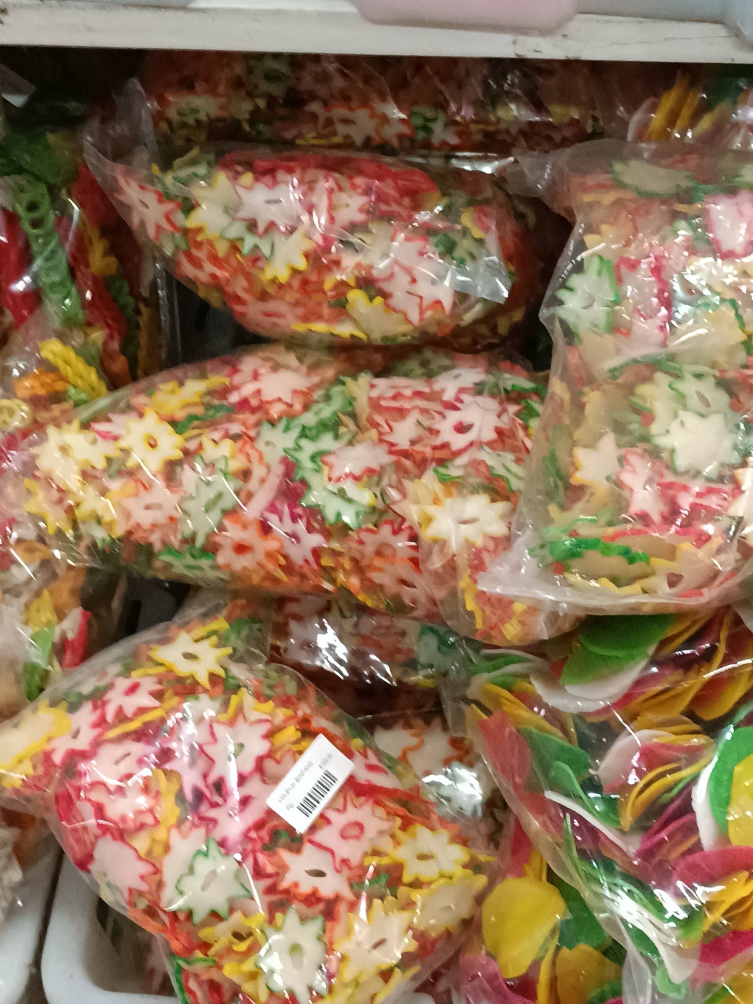 The image shows a close-up of several transparent plastic bags filled with colorful, star-shaped crackers. The crackers are in various shades of red, yellow, green, and white. They appear to be piled loosely within the bags, suggesting they are a bulk food item. In the bottom right of the image, a label is partially visible on one of the bags, with what looks like a barcode and some text in an Asian language. The bags are stacked on what appears to be a shelf, possibly in a store or market stall. The lighting is slightly harsh, creating reflections on the plastic packaging. There are no people visible in the image, and the focus is entirely on the food items. The background is blurred, but suggests an indoor environment with other items also possibly for sale. The specific location in Jetis, Indonesia is not evident from the image itself, but it is consistent with a scene one might find in a local market or a small grocery store there.