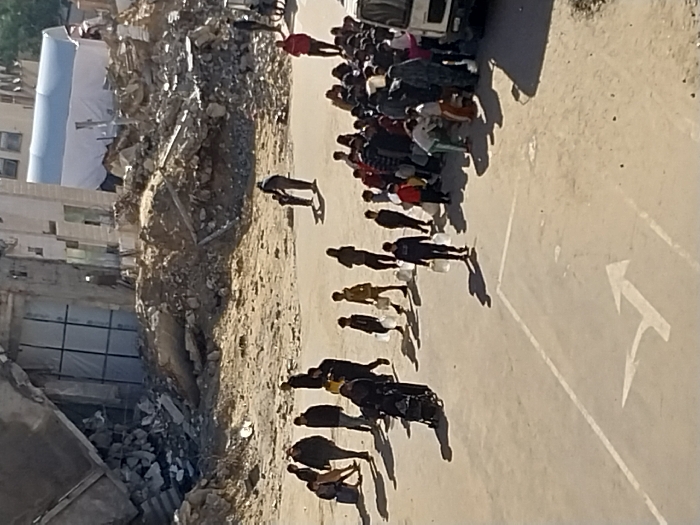The image depicts an outdoor scene in Khan Yunis, Palestinian Territory, characterized by urban devastation and civilian movement. In the background, the remains of multi-story buildings show extensive damage, with large piles of concrete rubble and exposed rebar. A large blue and white tent structure is visible behind the debris field on the right. The mid-ground consists of uneven, dusty ground. The foreground is a paved surface with a white painted arrow indicating a directional turn to the right.
Numerous individuals, including adults and children, are present. On the left, a group of approximately six people walks from left to right; one individual pushes a dark-colored stroller or cart, and several carry white plastic containers. Further in the mid-ground, a person holds the hand of a small child, walking away from the viewer towards the rubble. On the right, a larger crowd of over twenty individuals is gathered around a white utility vehicle, appearing to be a truck or van. Many in this crowd are holding white plastic containers or bags, and some are interacting with the vehicle, suggesting an aid or water distribution point. Other individuals are seen walking from right to left, also carrying white containers. Long shadows are cast across the paved ground from the individuals, indicating direct sunlight.