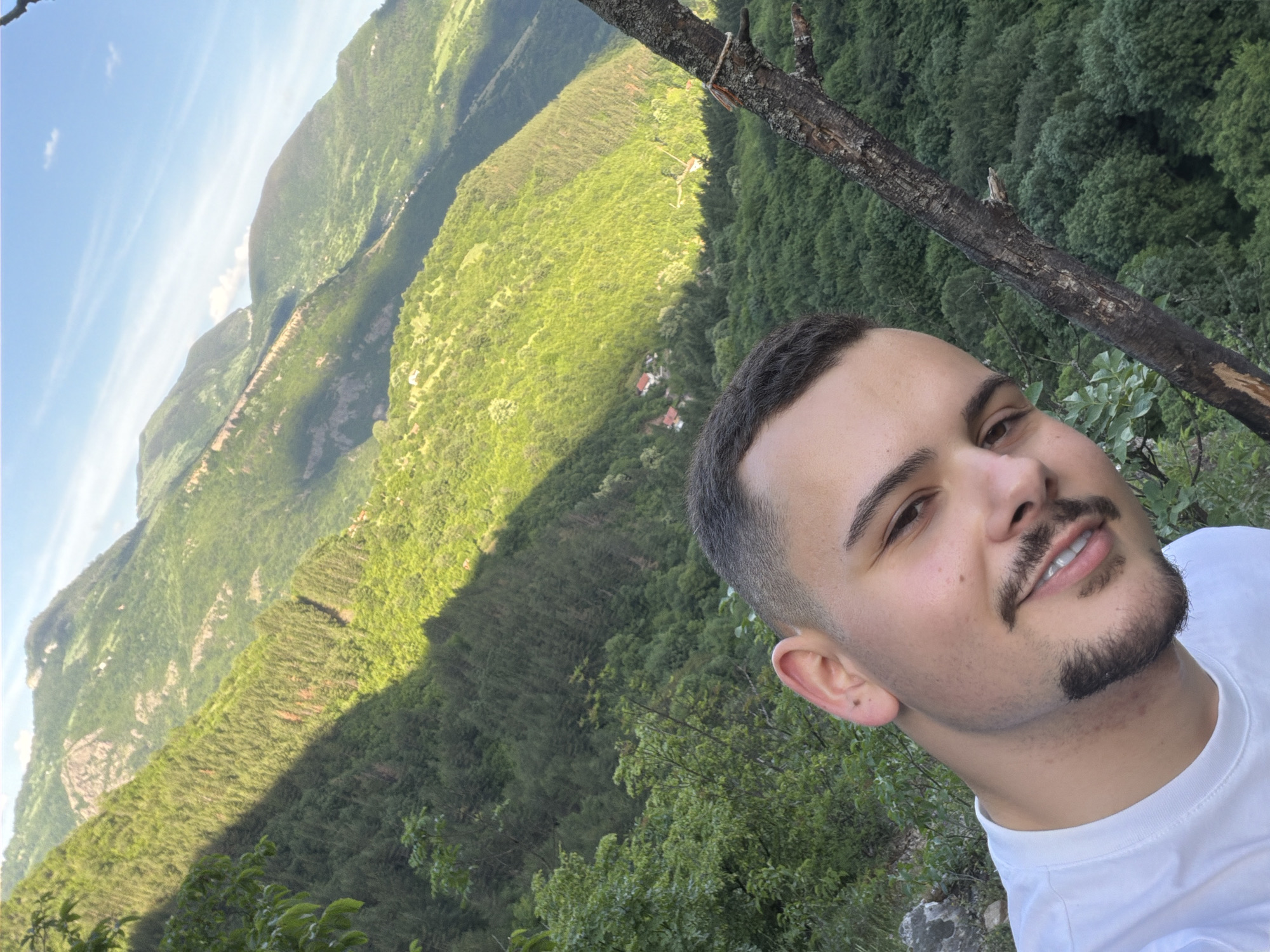 A selfie of a young man with light skin, short dark hair, and a dark mustache and beard, wearing a white T-shirt. He is positioned to the right of the frame. The background is a mountainous landscape densely covered with green trees, typical of a wooded area. A single, charred branch diagonally crosses the upper right portion of the frame, partially obscuring the background. The left side of the frame showcases a valley with varying shades of green vegetation. The sky is visible in the upper left corner, revealing a clear blue expanse with a few scattered clouds. Based on location information, the image likely depicts a setting in or near Zasele, Bulgaria, given the area's geography and environmental attributes.