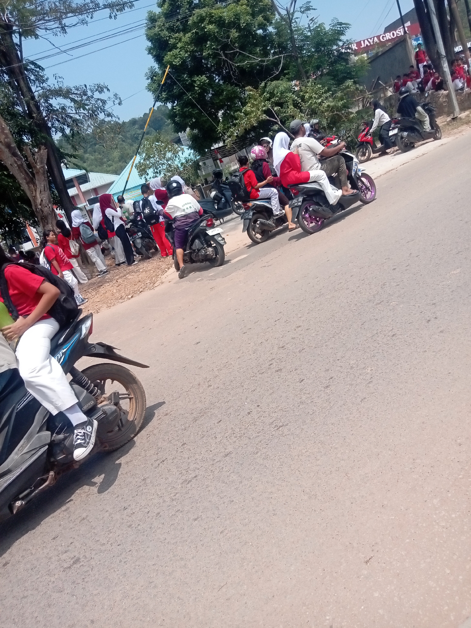 The image captures a street scene in Batam, Indonesia, likely near a school or educational institution given the presence of uniformed students. Several students in red and white uniforms are seen on and around motorcycles, suggesting a mode of transport or a gathering. One student in the foreground is riding a black and blue motorcycle, wearing white pants, a red shirt, and black Converse shoes. Another student on the same motorcycle is wearing a white hijab and a red shirt. In the background, more students are milling around, some on motorcycles, while others are standing or sitting on a low wall. A building with a sign "AR JAYA GROSIR" is visible, indicating a commercial establishment. The environment is characterized by a paved road, trees, and a bright, clear sky. The overall impression is one of everyday activity in a semi-urban Indonesian setting.