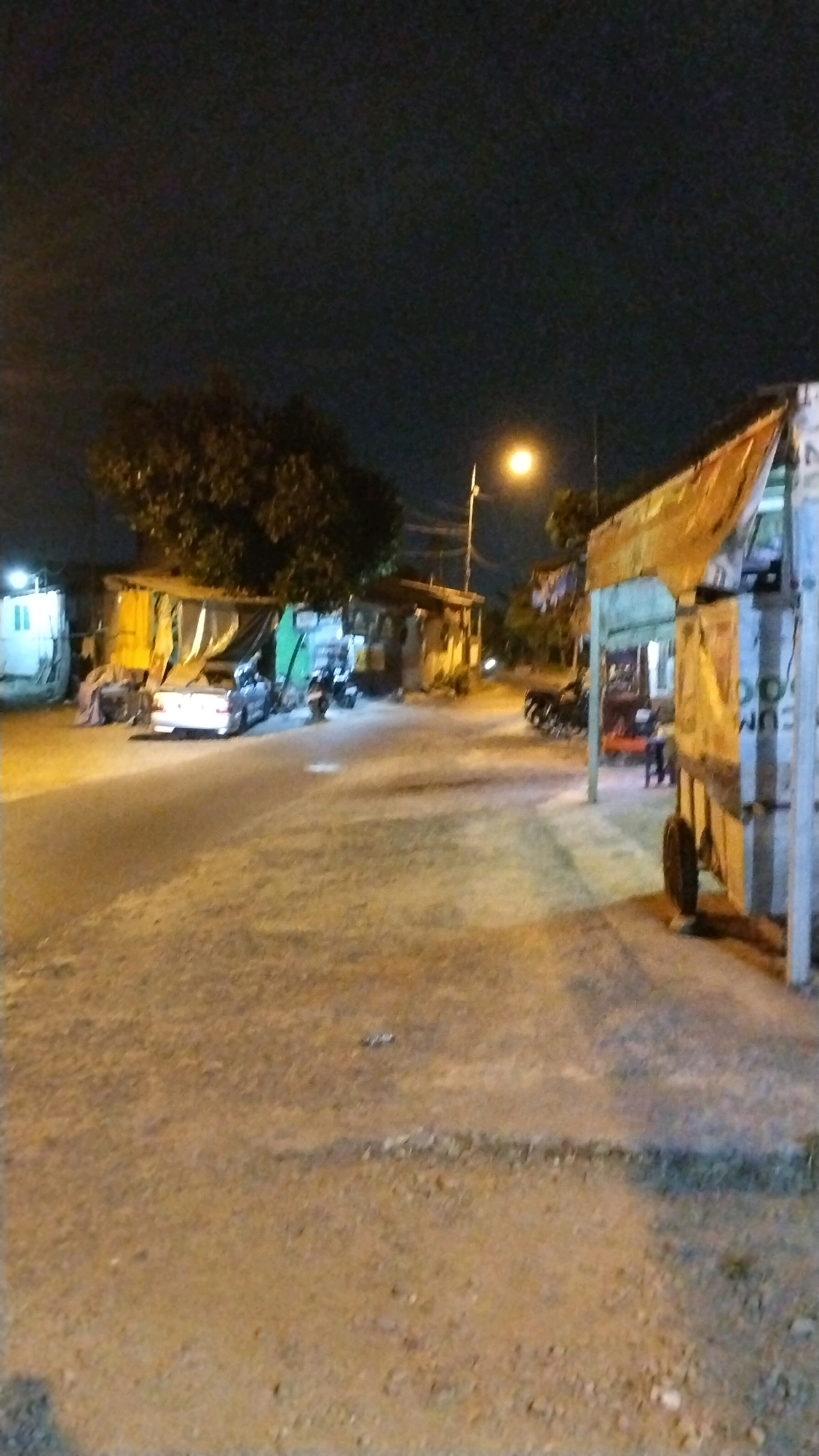 The image depicts a dimly lit street scene at night in Balongmojo, Indonesia. A paved road, with patches of gravel and dirt, runs through the foreground and extends into the middle ground. On the left side of the road, there are several small, makeshift structures or stalls with yellow and green tarps. A white vehicle, possibly a van or truck, is parked to the far left, partially obscured. A silver sedan is parked in front of one of the stalls, facing away from the camera. Further down the street, more stalls are visible, some illuminated by artificial light. On the right side of the road, a larger, more permanent-looking structure with a corrugated metal roof is situated. A cart with a wheel is positioned in front of this structure. A street lamp with a bright yellow light illuminates the scene from the upper middle. Trees with dense foliage are visible behind the stalls on the left. A few motorcycles are parked near the stalls on the left. The overall impression is of a commercial or residential area at night, likely a roadside market or a cluster of small businesses.
