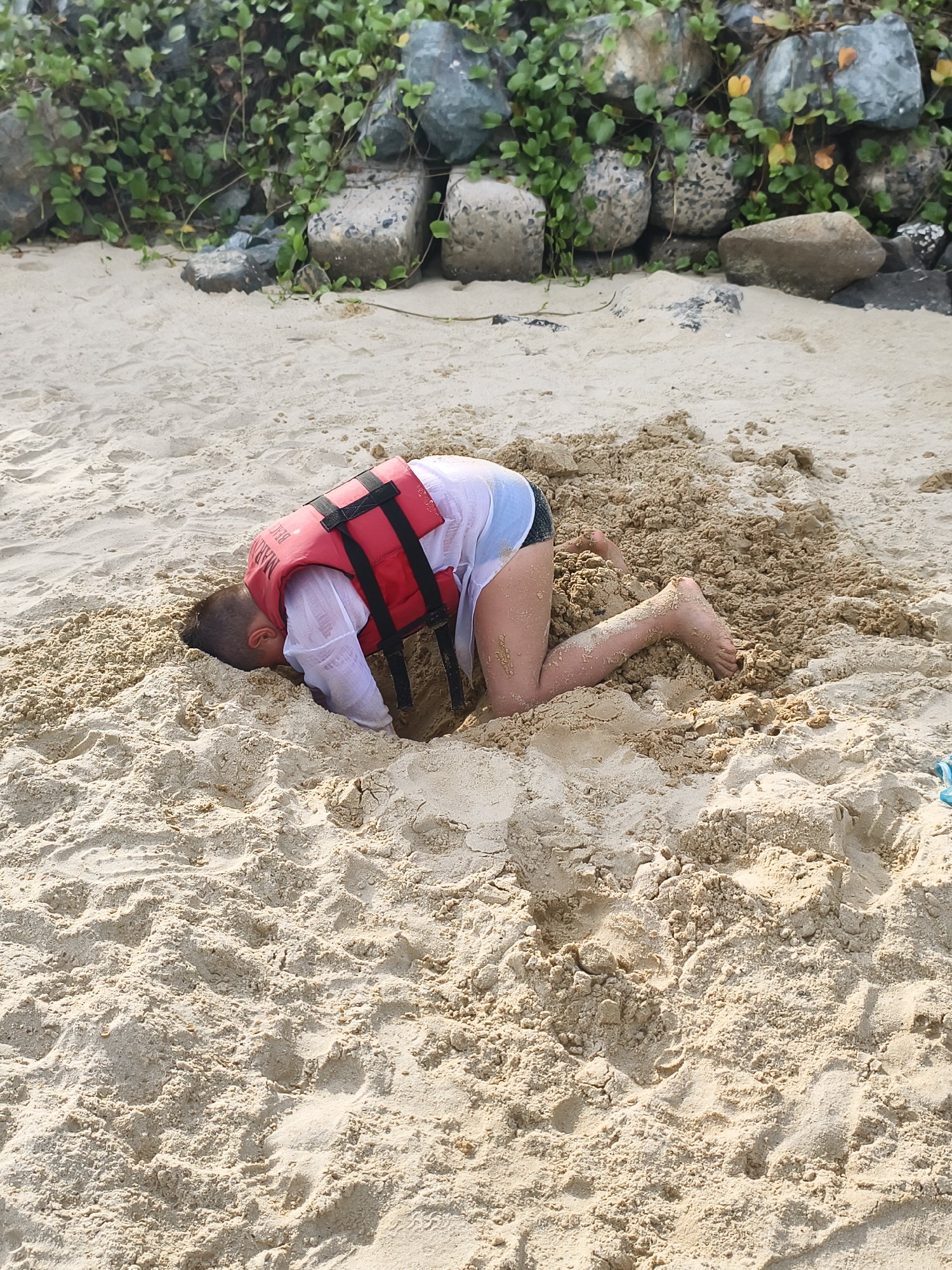 A child wearing a red life vest and a white shirt is playing in the sand on a beach. The child's head is buried in a hole dug into the sand. Their legs are visible, and they are kneeling in the sand. The background of the image shows a rocky area with green vegetation, likely bordering the beach. The sand is light brown and appears to be wet in some areas. The scene is outdoors, under daylight. The specific location within Bac Nha Trang Ward, Vietnam, cannot be determined from the image alone.
