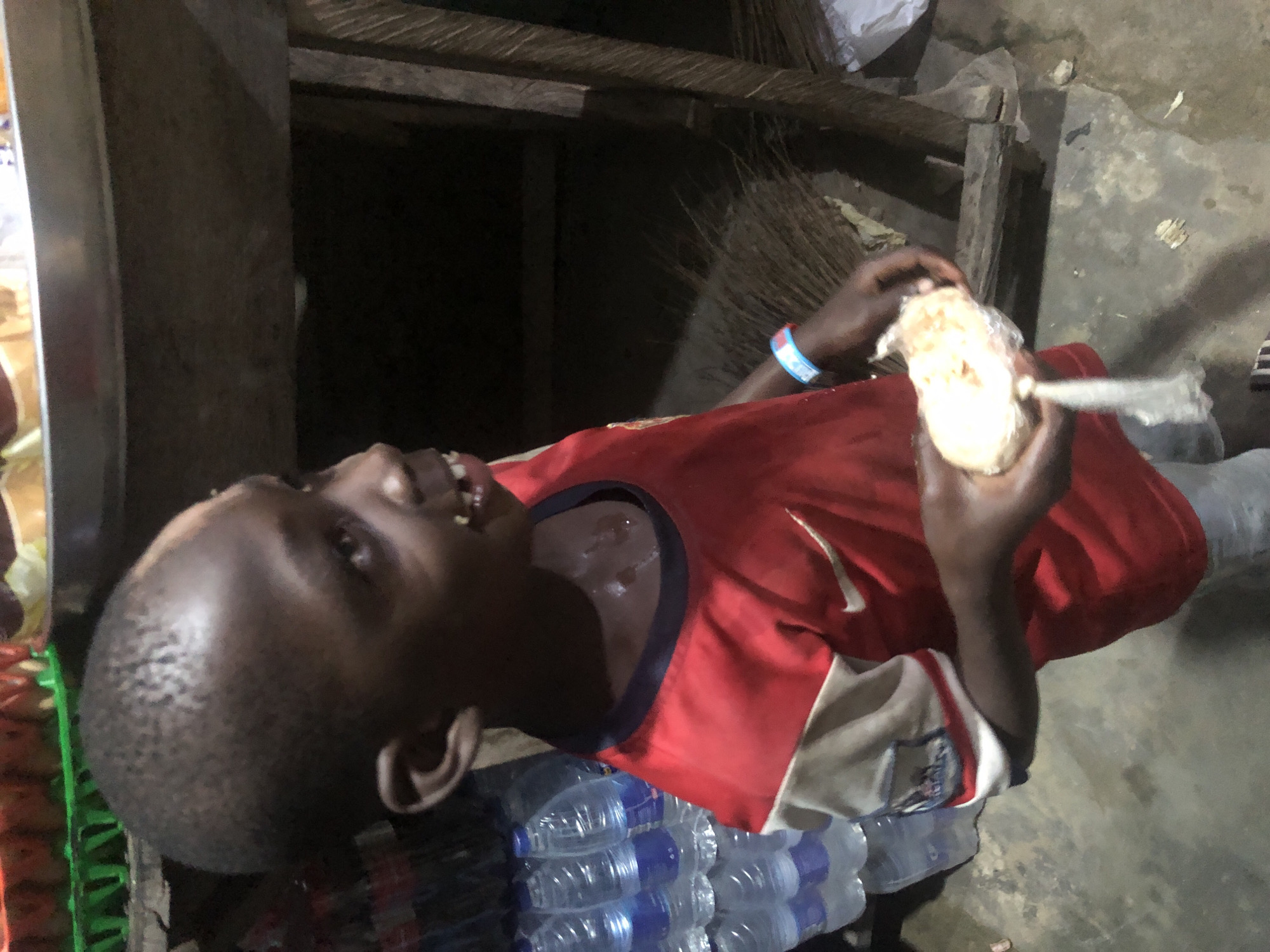 In Ado, Nigeria, a young boy wearing a red and white t-shirt and grey pants stands in front of a rustic wooden structure. He holds a piece of food wrapped in clear plastic in his hands. Behind him, several bottles of water are stacked. The ground is made of concrete, and straw-like material is visible on the right side of the image. The boy's head is shaved, and he has a small wound on his forehead. A colorful wristband is visible on his left wrist.