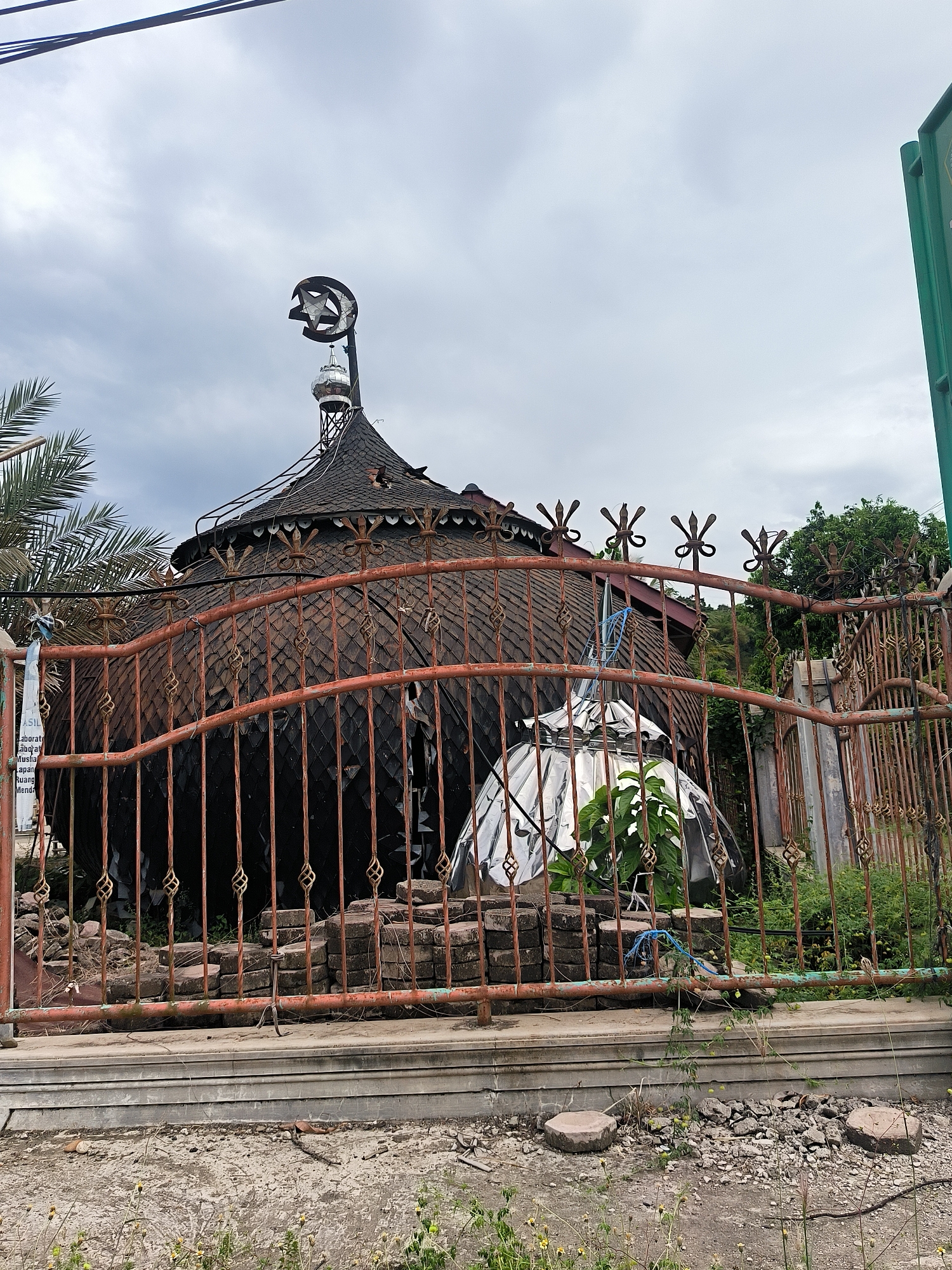 The image depicts a unique, circular building, likely a mosque or prayer hall, situated in Kuta Pungwa, Indonesia, under an overcast sky during daytime. The building features a dark, rounded roof covered in a pattern of small, overlapping shingles, giving it a scaled appearance. A tall, slender structure resembling a minaret rises from the center of the roof, topped with a prominent crescent moon and star finial, a symbol of Islam.
In the foreground, a heavily rusted, ornate metal fence spans horizontally across the frame, partially obscuring the lower section of the building. The fence has decorative spiral and star-shaped elements. Behind this fence, to the right of the main building, is a smaller, shiny, reflective dome-like structure, possibly a planter cover, with green plants growing inside. Piles of bricks or pavers are visible behind the fence.
To the left, palm fronds are visible. The ground in the immediate foreground consists of concrete and dirt with some weeds and scattered debris. Utility lines stretch across the upper left portion of the sky, and a partial green signpost is visible on the far right. A tattered white banner or sign hangs from the fence on the left, displaying partial Indonesian text including "...ASIL," "Laborat...", "budaya," "Musholla," "lapang...", and "Menda...". The scene conveys a rustic and somewhat weathered atmosphere.