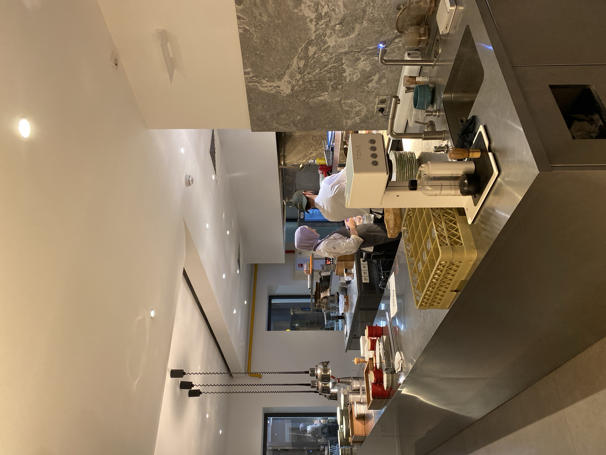 The image captures a commercial kitchen setting in Moscow, Russia. The location is identifiable from the "GENERATOR" sign written in Cyrillic. Two individuals are present, both seemingly kitchen staff, possibly preparing food or drinks. One wears a white chef's coat and a green cap, while the other is in a white uniform and a lilac hijab. They are positioned behind a metal counter, which features a sink area with faucets, various utensils, and a "TONE" branded appliance that might be a food or beverage preparation machine. A yellow plastic crate is also visible. Behind them, there's a window, likely leading to an outside view of the city. Above, the ceiling is white with embedded lights and hanging light fixtures. The walls are a mix of white and gray marble.