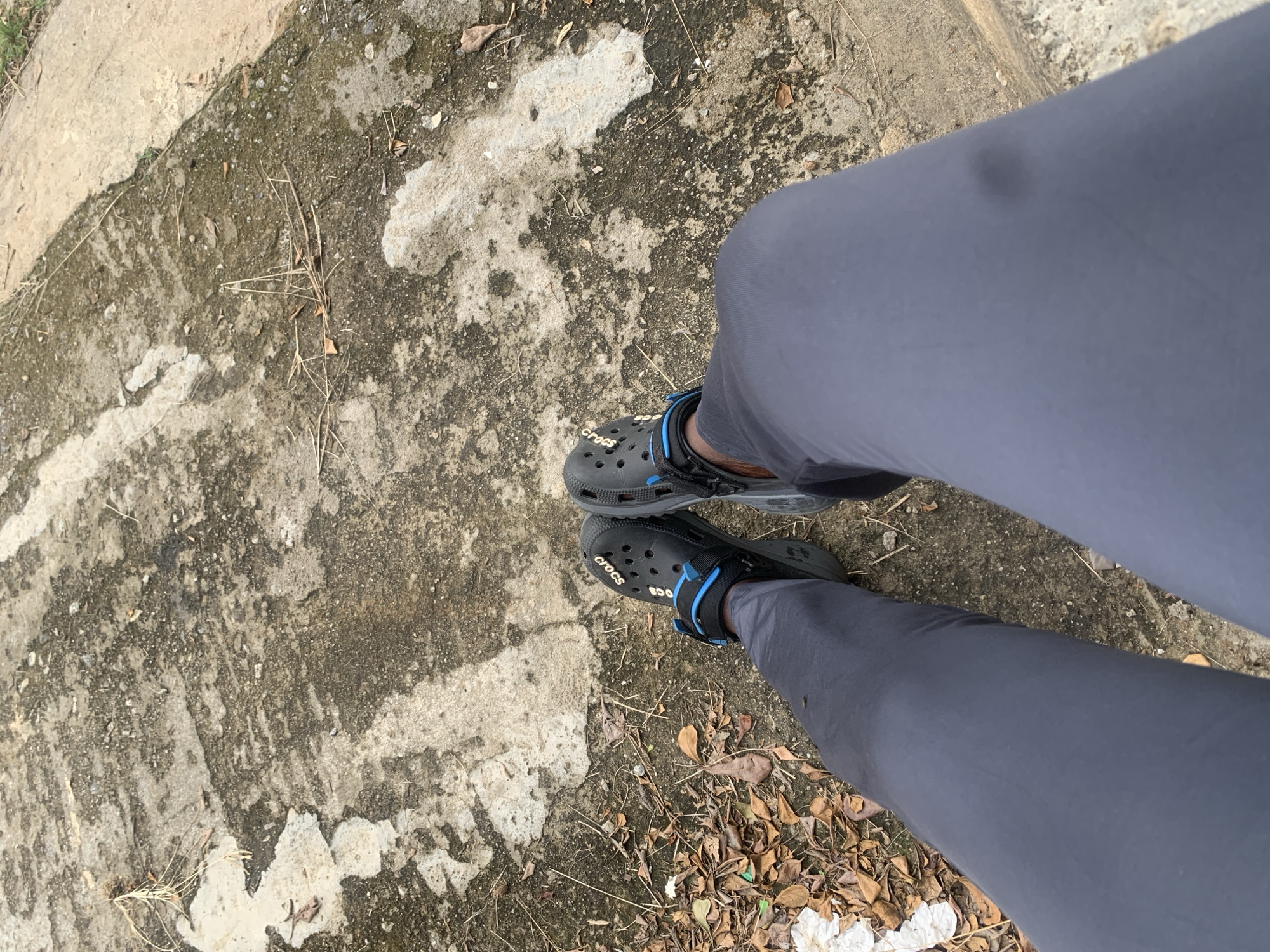The image shows a person's legs and feet wearing gray pants and black Crocs with blue accents, resting on a textured concrete surface. The surface appears weathered and is scattered with dry leaves and twigs. The scene is shot from a high-angle perspective, looking down at the subject's lower body. The concrete has patches of discoloration and a slightly damp appearance in some areas. The environment suggests an outdoor setting, possibly a pathway or walkway. The image does not provide any specific visual cues to pinpoint the location to Abuja, Nigeria.