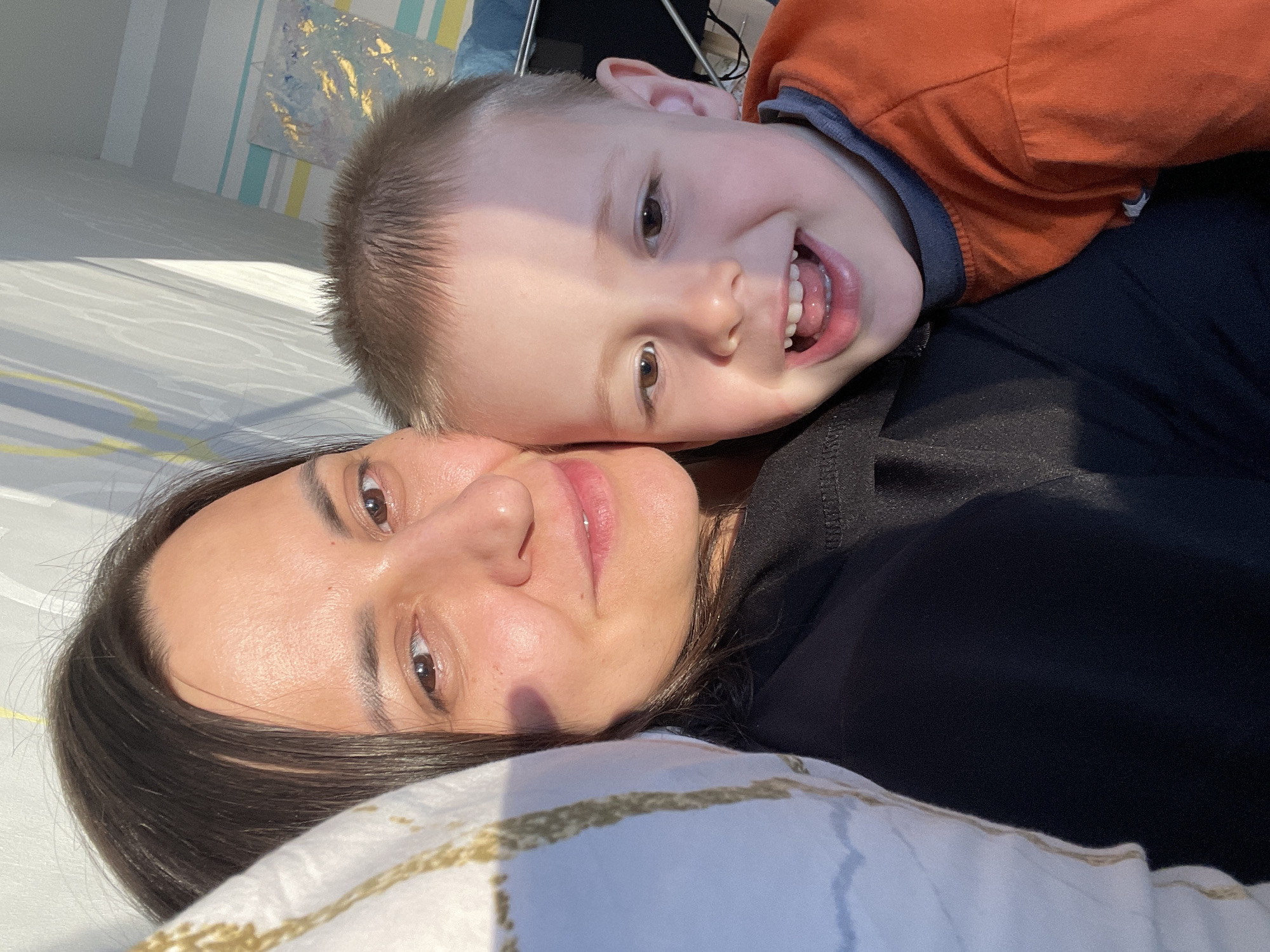 A close-up, top-down selfie shows a woman and a young boy lying down. The woman, in the foreground, has dark hair and is looking up at the camera with a slight smile. Her eyes are brown, and she is wearing a black shirt. The boy, who appears to be nuzzling into the woman's face, is wearing an orange shirt. He has light brown hair and a wide, joyful grin, showing his teeth and tongue. The background includes a striped wall, possibly in a bedroom, with some abstract art. The lighting suggests it is daytime, with sunlight casting shadows. The woman is resting her head on a pillow with a white and gold striped pattern. The overall mood is affectionate and happy, with the boy's ecstatic expression and close proximity to the woman. There is no visible text in the image. The location context is Izhevsk, Russia.