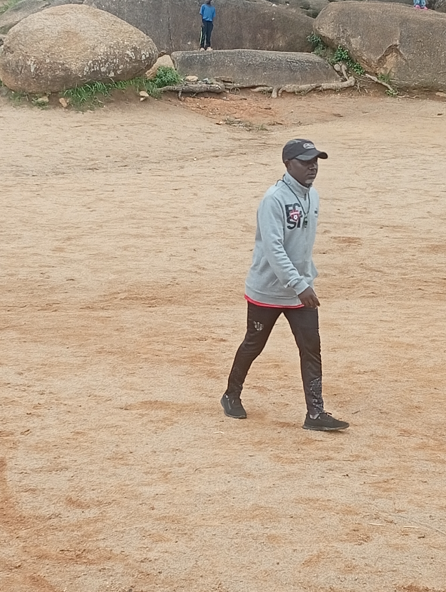 A medium shot shows a man walking across a sandy, outdoor area with large rocks in the background. The man is wearing a light gray hoodie with red trim at the hem and wrists, black pants with a graphic design on the lower leg, a black baseball cap, and black athletic shoes. He is walking from left to right across the frame, with his head turned slightly to his left. In the background, a person in a blue shirt and black pants stands on top of a large rock formation. The environment appears to be a natural, rocky landscape, possibly in the city of Jos, Nigeria, known for its granite outcrops. The ground is covered in a light brown sand, with some sparse vegetation growing around the base of the rocks. The sky is not visible in the frame.