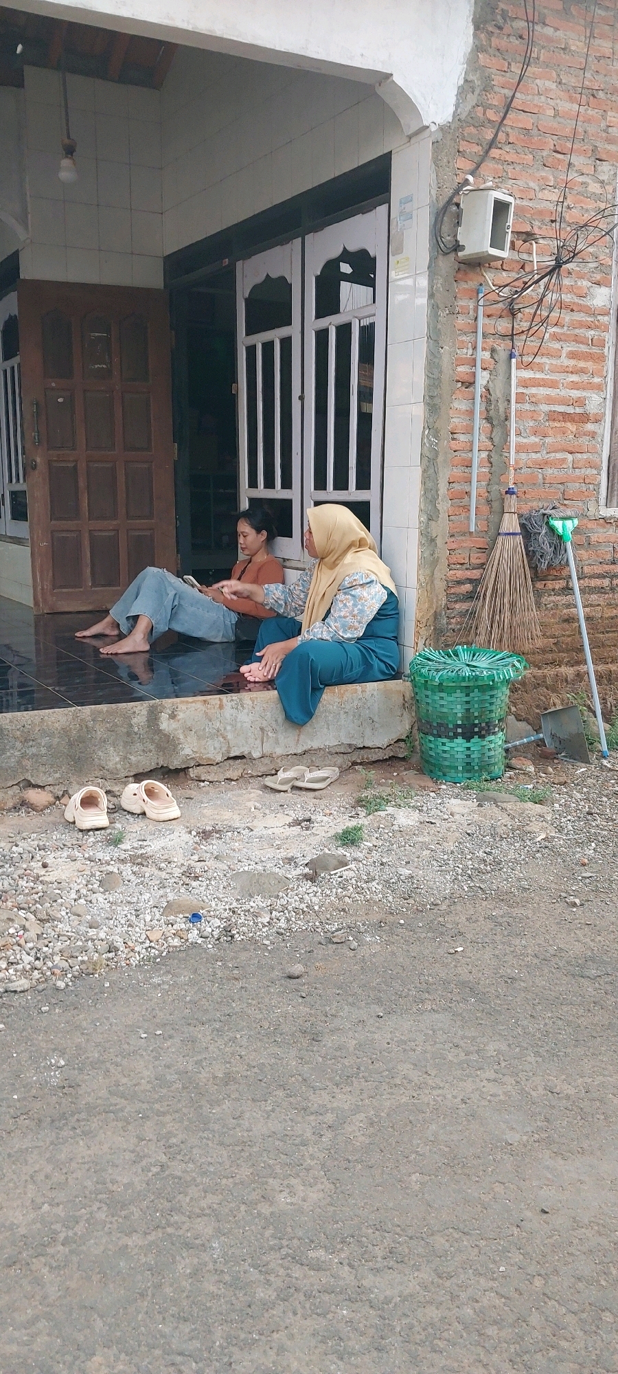 Two individuals are seated on the concrete step in front of a building. The individual on the left is a young woman with dark hair pulled back, wearing a rust-colored long-sleeved shirt and loose-fitting blue jeans. She is sitting cross-legged with her bare feet visible, and appears to be looking at a phone or tablet in her lap. The individual on the right is an older woman wearing a tan hijab and a patterned blue and white long-sleeved top with teal pants. She is seated with her legs extended, also with bare feet visible. She is gesturing with her right hand and looking towards the left.
The building behind them has a wooden door on the left and white-framed glass double doors on the right. The wall is constructed of red brick. To the right of the step, a broom and dustpan are leaning against the wall, and a green woven basket is situated on the ground. Several pairs of sandals are placed on the gravelly ground in front of the step. The scene appears to be an outdoor area in front of a residence, possibly in Keling, Indonesia. The ground in the foreground is a paved road or pathway with loose gravel.