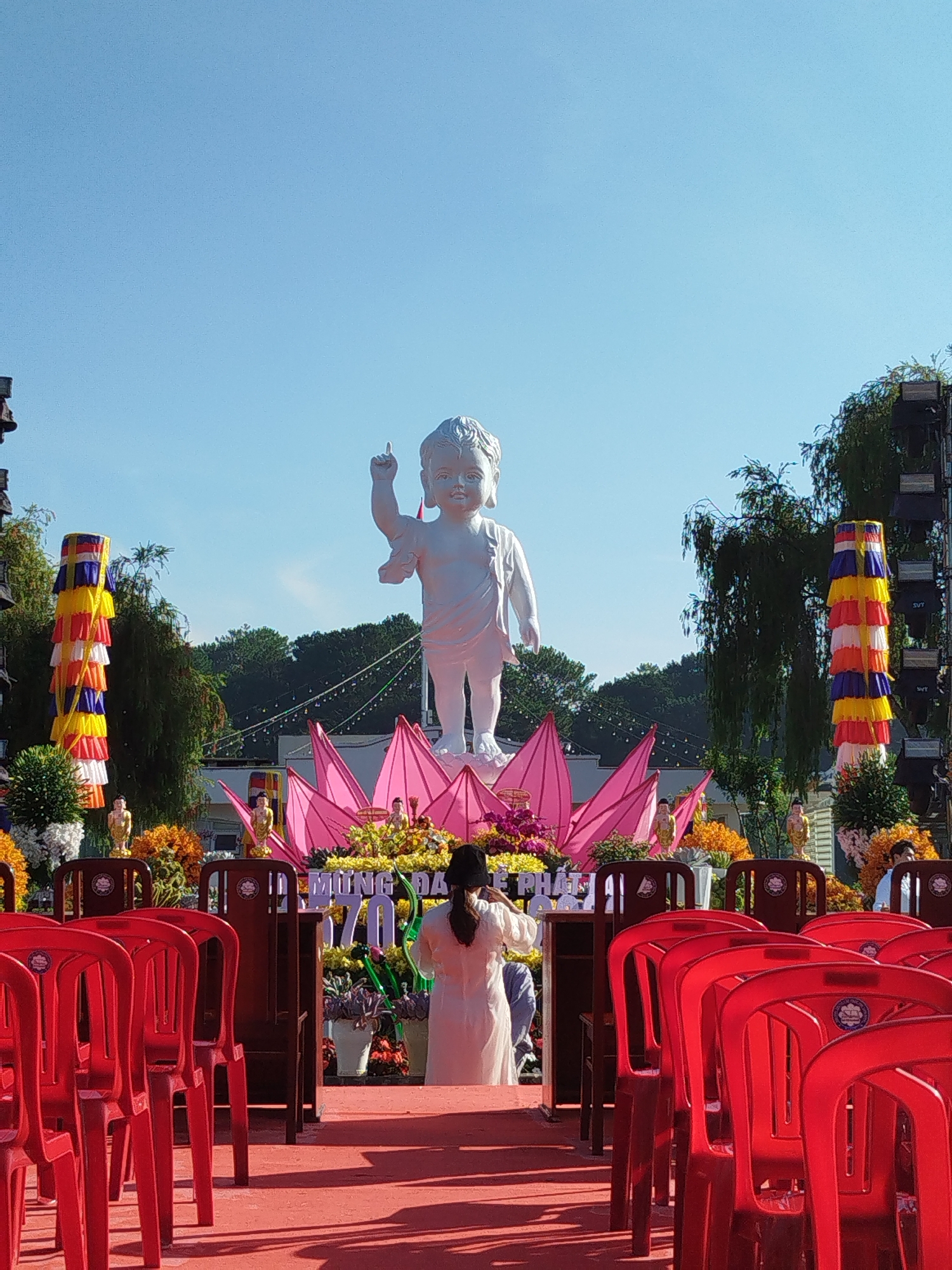 A daytime shot displays a statue of the Buddha as a baby, standing on a pink lotus flower. A woman in a white robe stands in front of the statue, appearing to be in prayer. Rows of red plastic chairs line the foreground, and the ground is covered with red fabric. The event is set up with colorful banners and flower arrangements. Trees and buildings are visible in the background under a clear blue sky. The image appears to be captured at a Buddhist temple or location in Da Lat, Vietnam.