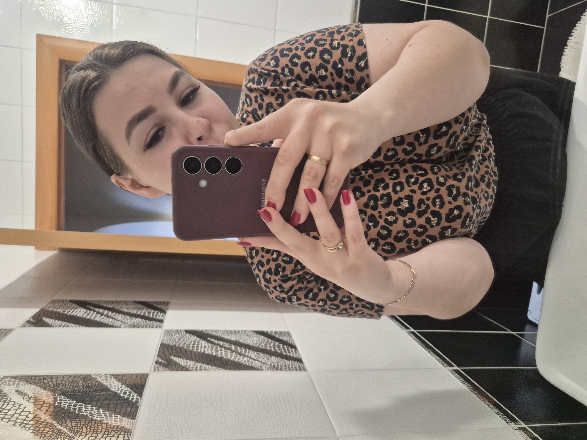 A fair-skinned female is taking a selfie in a bathroom. She is holding a maroon smartphone with three camera lenses up to her face, partially obscuring her features. She has red nail polish and wears a gold ring on her ring finger, along with a gold bracelet on her wrist. She is wearing a leopard-print top and dark-colored bottoms. The bathroom is tiled with a pattern of white, black, and gray tiles with an animal print design. A framed picture or mirror is visible in the background. The location is potentially Tobolsk, Russia.