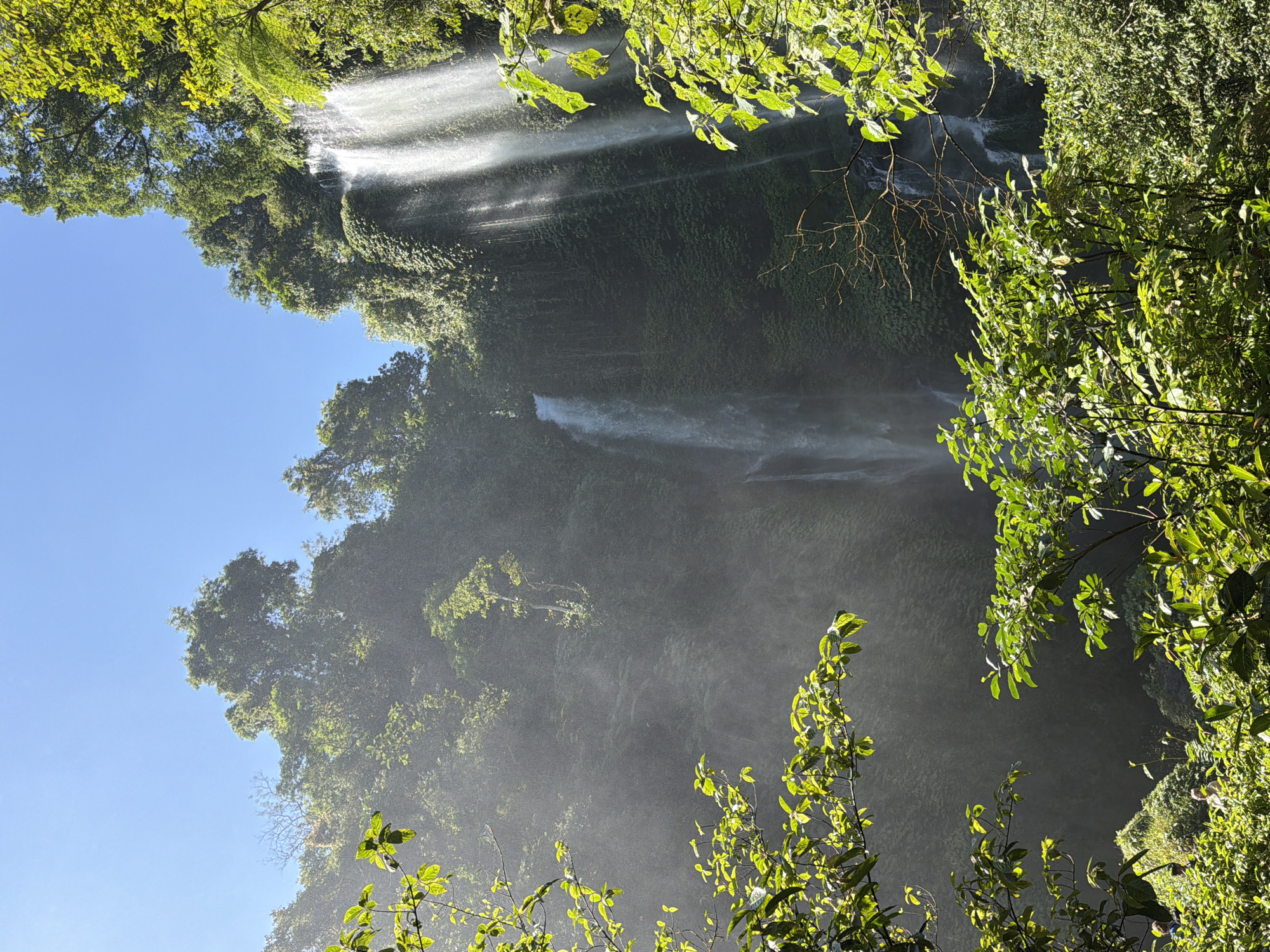 The image shows a tall, multi-tiered waterfall in a lush, tropical environment. The waterfall cascades down a dark, moss-covered cliff face, with mist rising from the impact of the water. Lush green foliage, including trees and vines, surrounds the waterfall, with sunlight filtering through the leaves and creating a hazy, ethereal atmosphere. In the foreground, the leaves of several trees are visible, illuminated by the sunlight. In the lower portion of the image, some people can be seen amidst the dense vegetation, appearing to be enjoying the natural scenery. The sky above is a clear blue. The scene is likely Sekumpul Waterfall, a prominent natural attraction located in Sawan, Buleleng Regency, Bali, Indonesia.