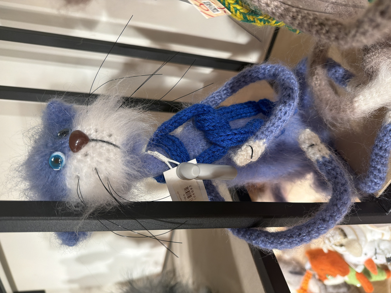 The image, taken in Saint Petersburg, Russia, features a fluffy, knitted or crocheted plush toy resembling a blue and white cat, displayed horizontally on a black metal shelf. The toy has a shaggy, fuzzy texture, predominantly in varying shades of light blue on its body and ears, contrasting with white fur on its face and chest. Its face is detailed with a shiny, bright blue eye on the left, a small brown nose, and several long, thin black whiskers extending from its white cheeks.
A dark blue knitted strap or handle is attached to the toy's back, suggesting it could be a small bag or a decorative item. A white tag, bearing a black barcode and numbers (partially visible as "006002" and "100"), is secured to the toy. In the blurred background, other knitted or textile items in various colors, including gray, green, and orange, are visible, implying a retail setting such as a craft store or market stall. The lighting appears to be artificial and bright. There are no people visible, and the primary activity is the display of the merchandise.