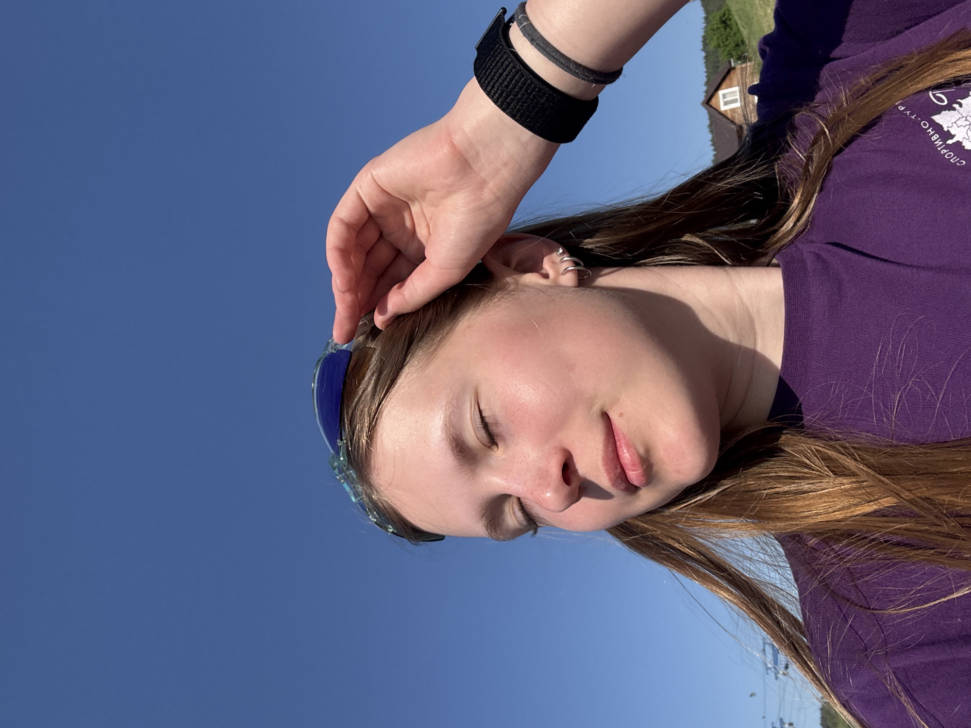 A person, presumably female, with closed eyes, fair skin, and light brown hair is centered in the image. They are wearing blue sunglasses and a purple t-shirt with Cyrillic text. A black watch or wristband is visible on their left wrist. Their left hand is touching their forehead. The background is a clear blue sky. A building and foliage are visible in the distance in the top right corner. Based on the Cyrillic text on the shirt, and the possible distant landscape, the location could be speculated to be Khvalynsk, Russia.