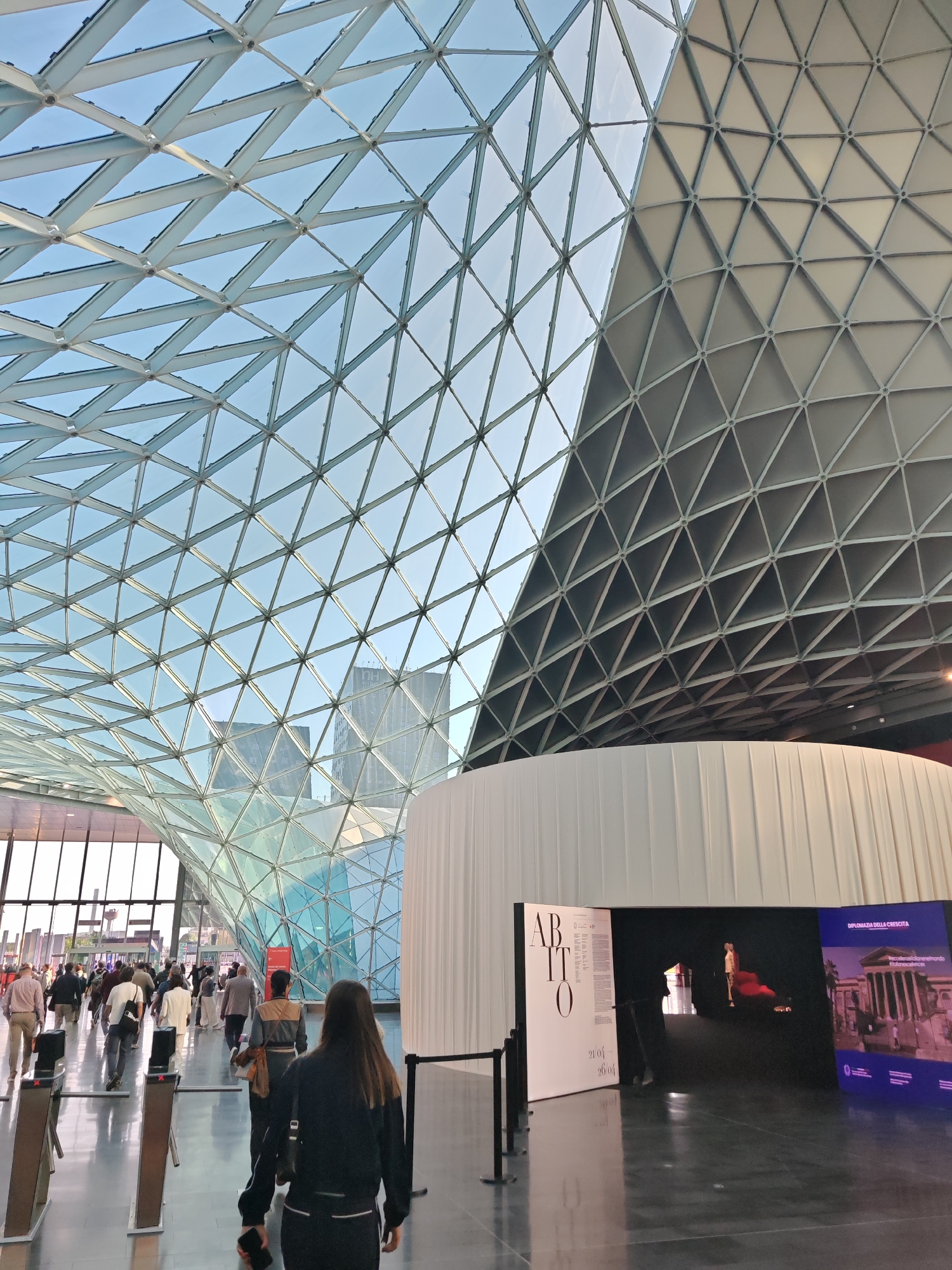 This media file captures the interior of a large, modern architectural space, likely an exhibition hall or convention center, characterized by its distinctive geometric glass and metal ceiling. The ceiling forms a complex pattern of triangles, allowing abundant natural light to fill the area, with a clear blue sky visible overhead. Part of the ceiling structure on the right is opaque, also featuring a similar triangular grid pattern.
In the foreground and mid-ground, a group of people is seen from behind, walking along a wide pathway, suggesting movement through an entrance or towards an event. They are dressed in various attires, from casual to business casual. Several dark, metallic turnstile-like barriers line the path.
On the right side of the image, a large, curved white structure serves as an exhibition booth or display area. A prominent display stand features the Italian word "ABITO" along with the dates "21.03 - 26.04", indicating an event or exhibition. Further within the dark opening of this structure, a screen displays a figure. To the far right, another blue screen shows the Italian text "DIPLOMAZIA DELLA CRESCITA" and an image of a classical building. Tall, modern city buildings are visible through the glass ceiling in the background. The scene depicts a bustling daytime environment under a clear sky.