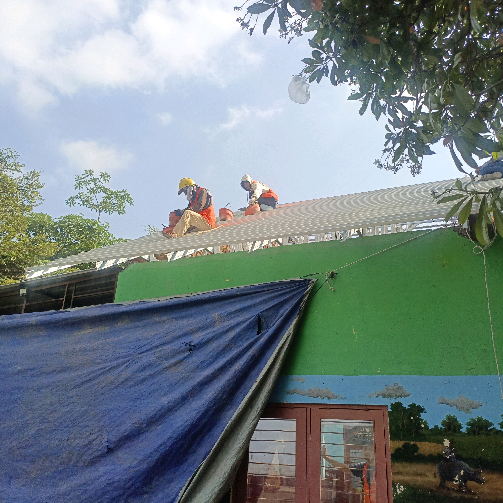 Two construction workers, wearing hard hats and safety vests, are working on the roof of a building in Salatiga, Indonesia. One worker is wearing a yellow hard hat and orange safety vest, and is seated, holding a tool and a red bucket. The other worker is wearing a white hoodie and orange safety vest, and is kneeling, also holding a tool and a red bucket. The roof appears to be made of metal sheeting supported by metal beams. A large blue tarp is draped over the lower part of the building, covering a mural that depicts a rural landscape with a person riding a water buffalo. Green trees and blue sky with clouds are visible in the background.
