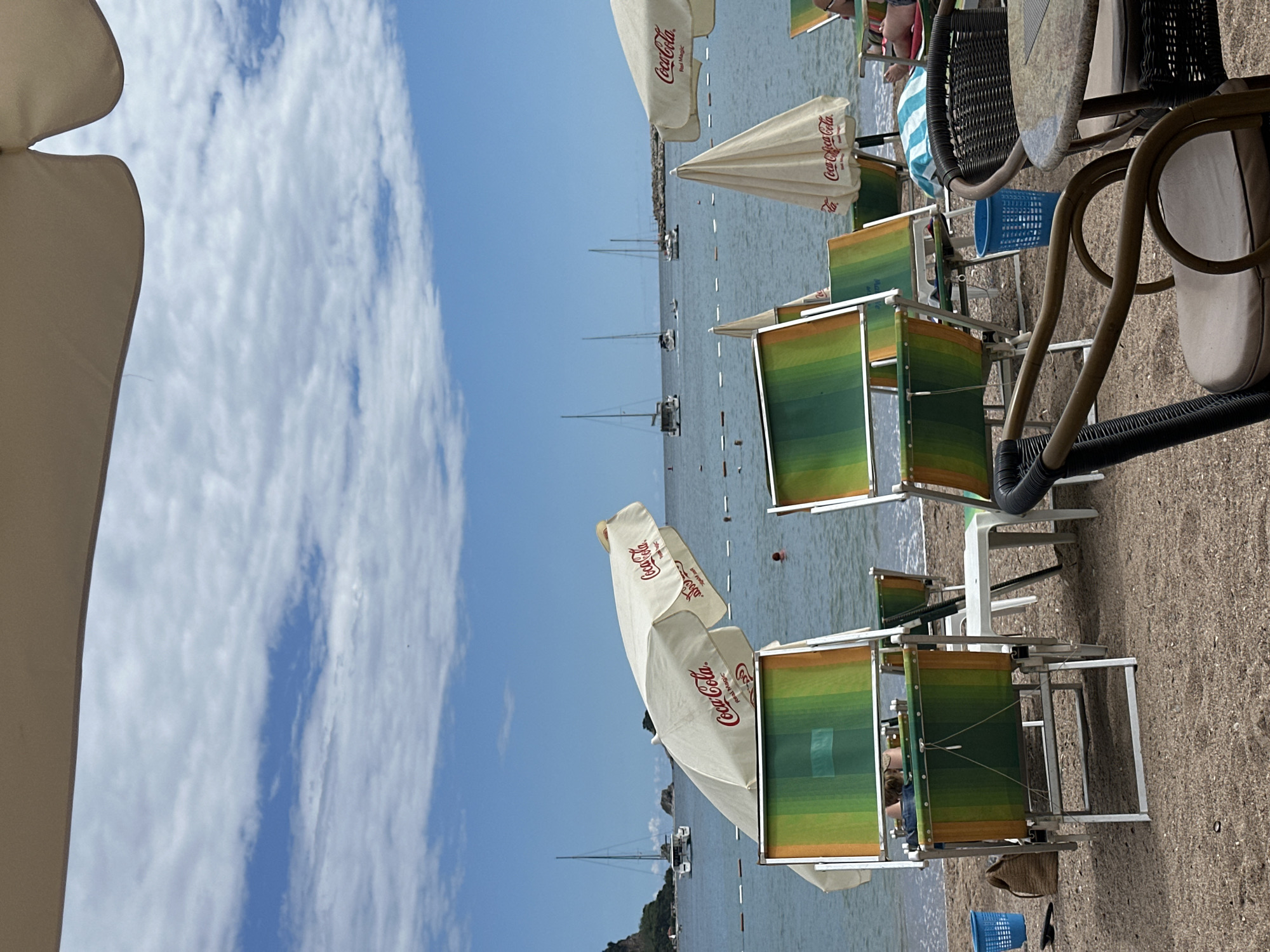 This image captures a bright, leisurely beach scene, appearing as if taken from under a sunshade on the shore, looking out towards the sea. The image, though rotated 90 degrees clockwise, depicts a typical beach setting under clear skies.
On the right side of the frame, the sandy beach is furnished with numerous cream-colored sun umbrellas, many prominently featuring the "Coca-Cola" logo in red, with one also displaying "Red Magic." Green and orange striped beach loungers are arranged on the sand, some appearing to be occupied by people partially visible, such as legs and a torso in a striped towel or shirt. Small tables are placed alongside some chairs, and a blue plastic basket and a woven bag are visible on the sand. Closer to the viewer on the far right, dark brown wicker-style chairs and a table suggest a cafe or an extended seating area.
The calm blue sea dominates the left and central parts of the image, with several sailboats and motorboats dotted across its surface, some appearing anchored in the distance. The horizon line shows a faint outline of distant land or hills. The upper portion of the image reveals a bright blue sky adorned with scattered, wispy white clouds.
The scene conveys a relaxed, sunny daytime atmosphere, typical of a holiday destination. The presence of beach furniture and boats indicates a popular coastal spot, consistent with a location like Budva, Montenegro. The visible text consists solely of "Coca-Cola" and "Red Magic" on the umbrellas.