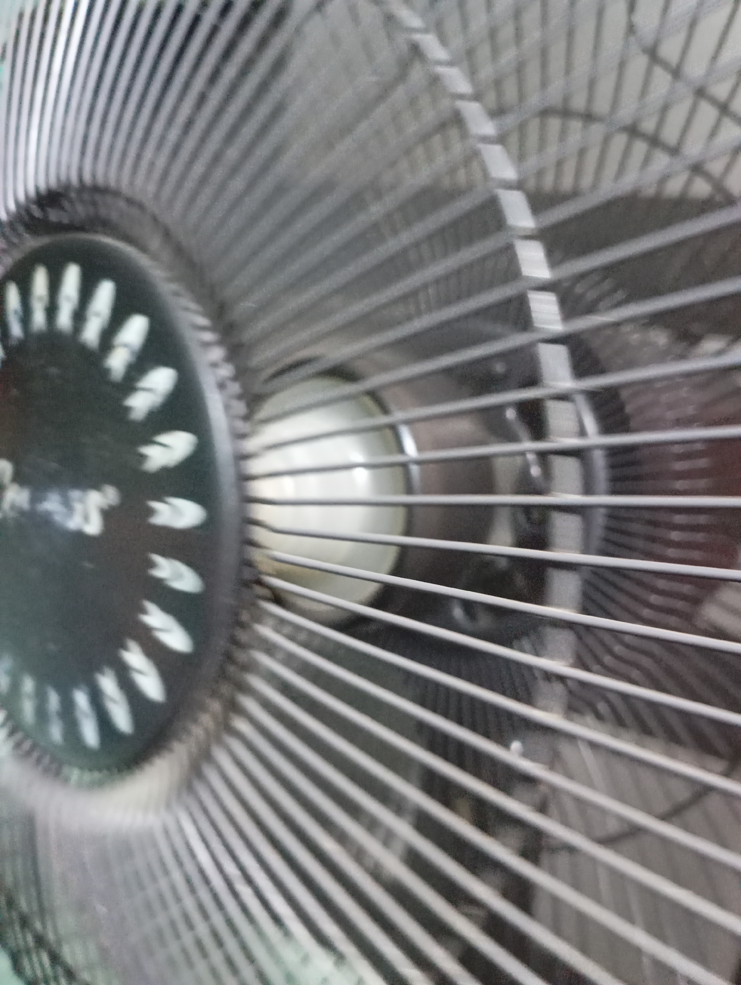 The image is a close-up, slightly blurry shot of a black electric fan's front grille and part of its motor housing. The fan grille consists of many thin, parallel metal bars. The motor housing has a circular black surface with a white logo or text, some of which is visible and appears to say "MAS" with some stylized markings around it. Behind the grille, a white, somewhat translucent circular object, likely the fan blades, is partially visible. The lighting suggests an indoor setting, possibly a room. There is no visible indication of the specific location within Kecamatan Haurgeulis, Indonesia, as the image focuses solely on the fan.