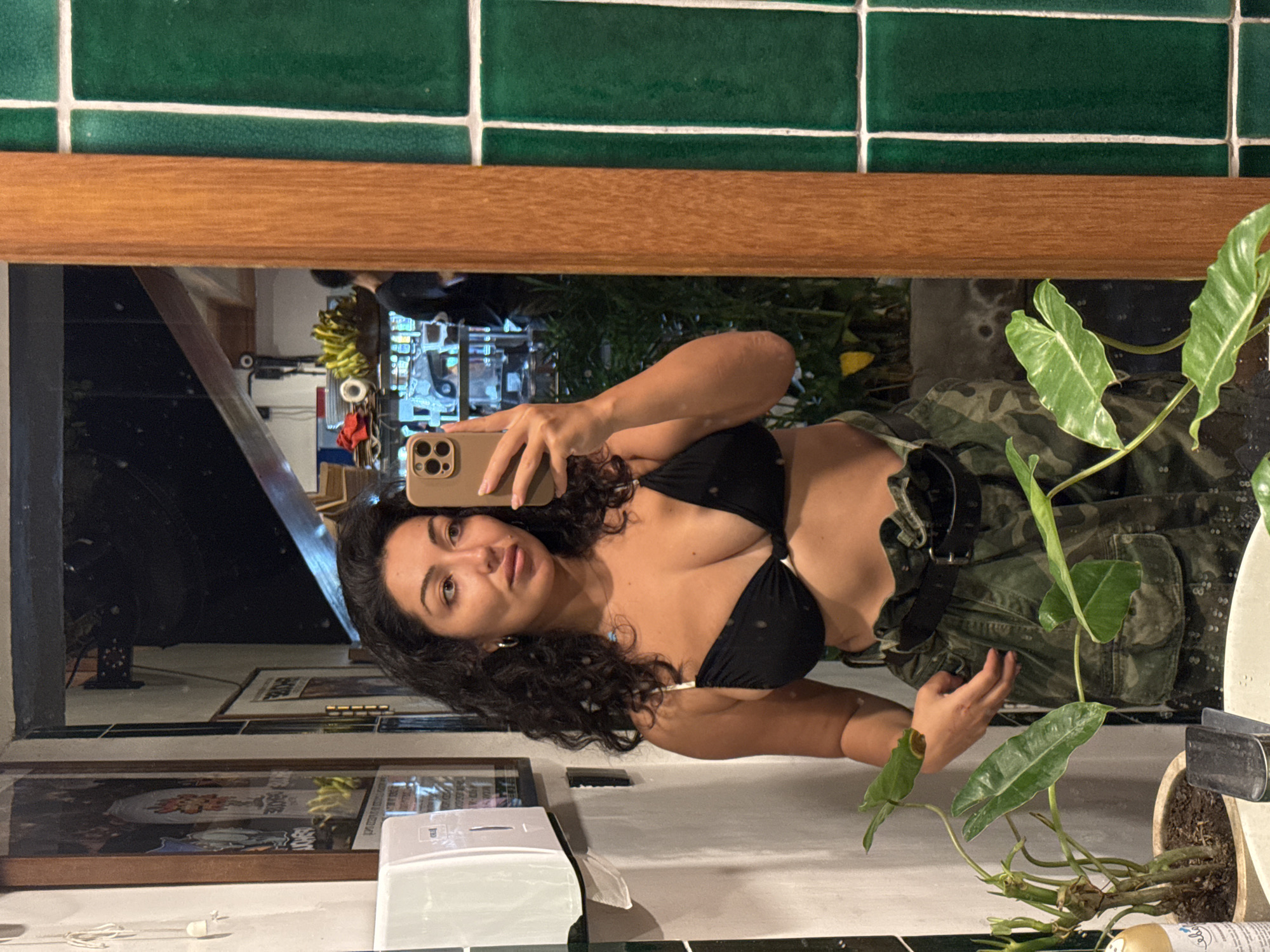 A woman with dark, curly hair is taking a mirror selfie in what appears to be a restroom. She is wearing a black bikini top and camouflage pants. In the background, a wooden beam divides the image, with green tiles on the right and a mirror reflecting a room with plants and a bar area. The location is identified as Mengwi, Indonesia.
